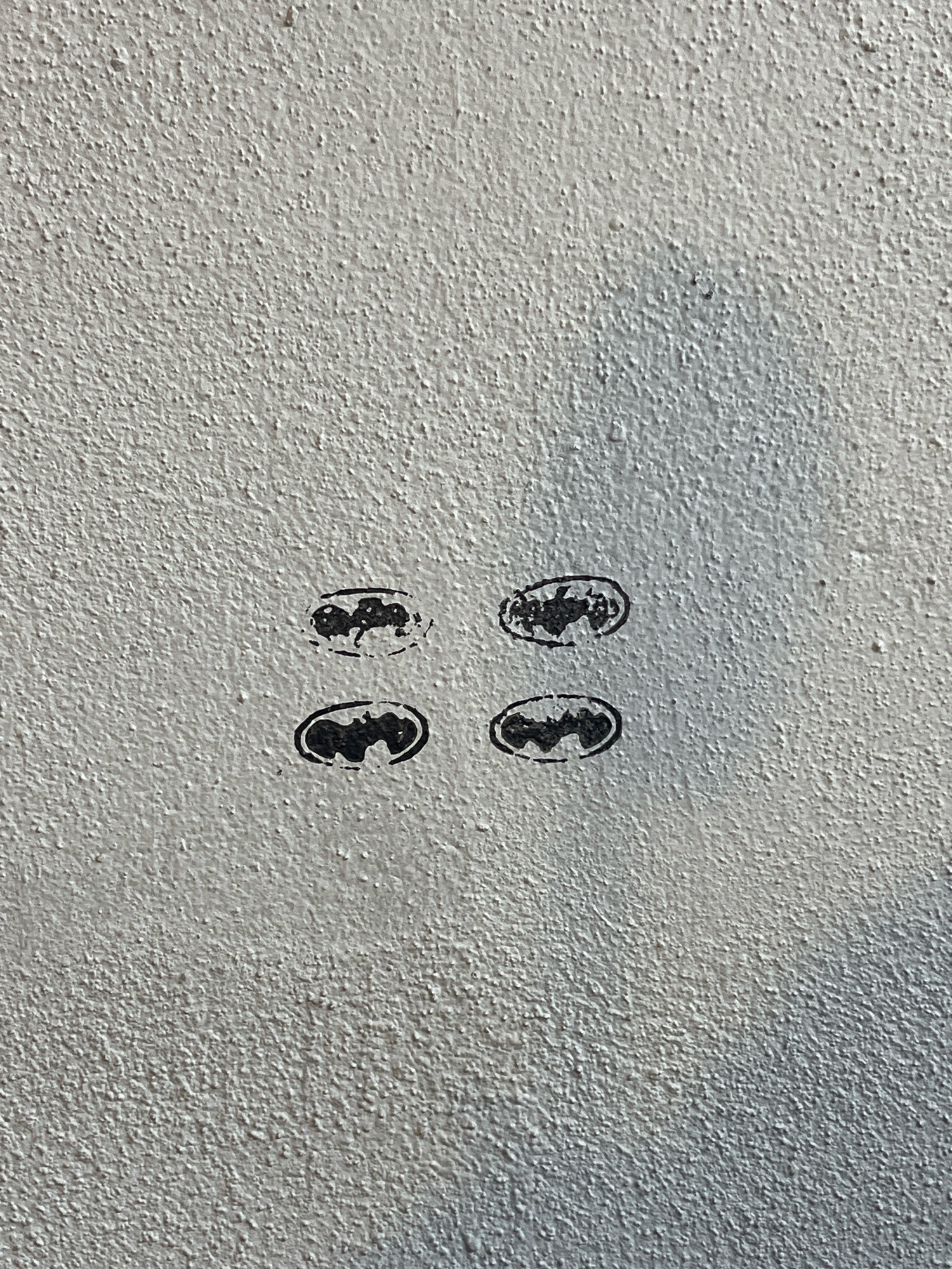 Four small, black bat symbols are stamped onto a textured, light-colored concrete wall. The symbols are arranged in a two-by-two grid formation. The wall appears to be an exterior surface, possibly part of a building or a barrier. The lighting suggests it is daytime, with shadows cast on the wall. There is no indication of the specific location in Kuala Lumpur, Malaysia, other than the presence of the wall itself. No individuals or actions are depicted.