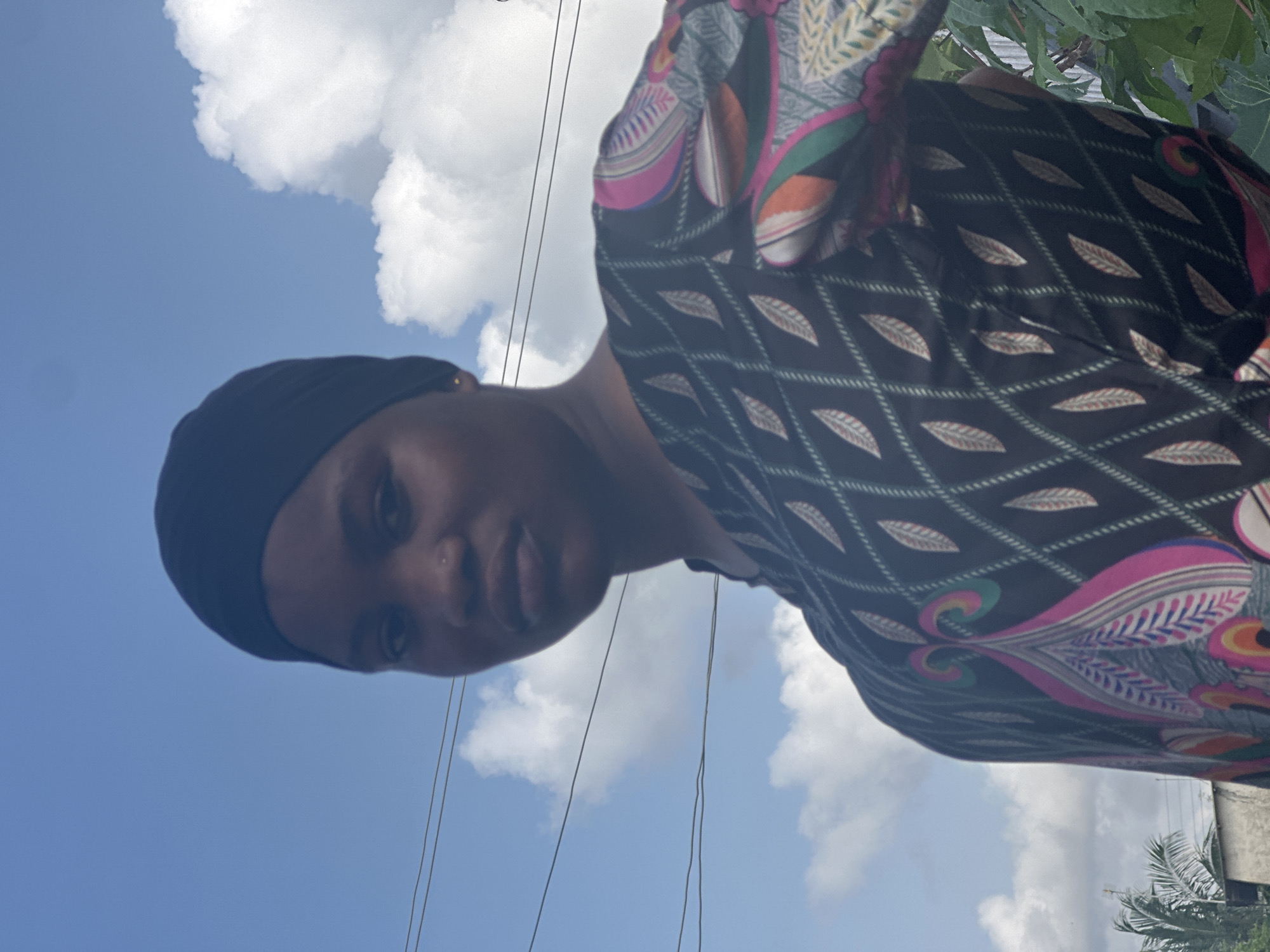 A young woman is featured in a portrait-style photograph, captured outdoors in bright daylight. The image is taken from a low-angle perspective, presenting her against a clear blue sky adorned with scattered white, fluffy clouds. She has a dark complexion and wears a plain black head covering, possibly a hijab or scarf, along with a patterned top. The top features a dark base, likely black, decorated with repeating light-colored, leaf-like patterns and sections of vibrant, intricate paisley designs in hues of pink, green, orange, and blue.
The woman looks directly at the camera with a neutral to slightly serious expression and has a nose piercing. In the background, thin electrical wires are visible crossing the sky. To the right, there is a hint of green foliage, possibly a palm tree, and the faint outline of a building structure. The overall lighting suggests it is daytime with abundant natural sunlight. The location context provided indicates this scene is in Badagry, Nigeria. There is no visible text in the image.