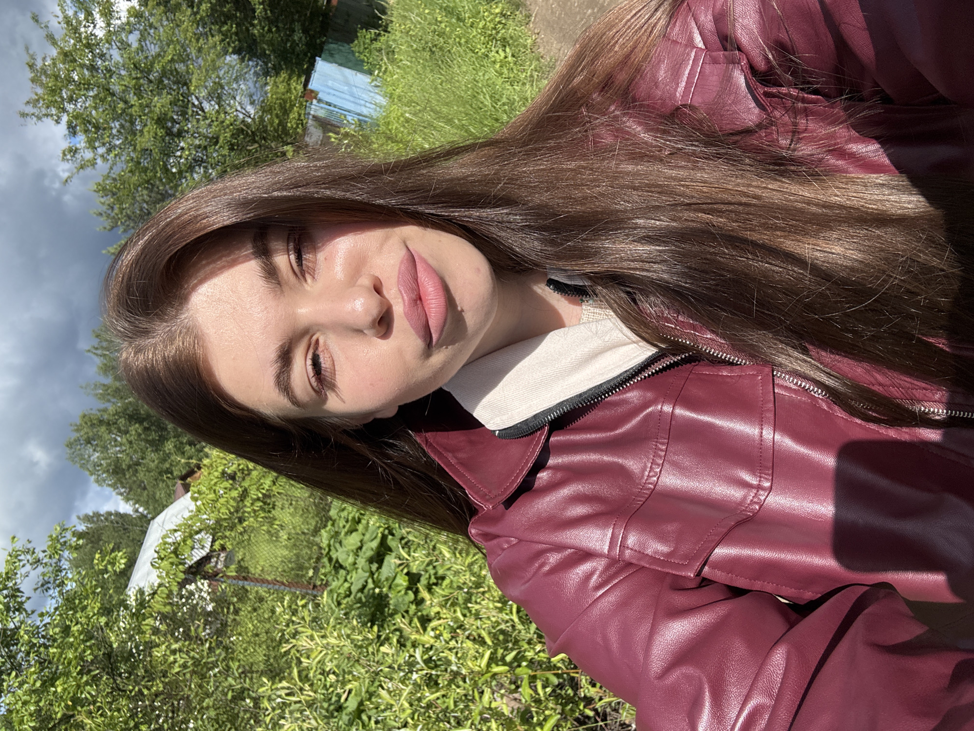 A woman with long brown hair is wearing a maroon leather jacket and a white shirt underneath. She is outdoors in a grassy area with trees and vegetation, potentially in a garden or park. In the background, there are hints of a structure, possibly a building or fence, partially obscured by the greenery. The photo appears to be taken in daylight, with some shadows indicating the presence of sunlight. Without more context, it's difficult to confirm the exact location as Шевлягино, Russia, or provide more specific details.