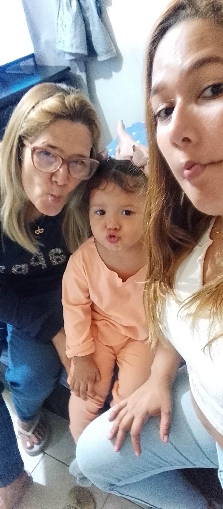 The image shows three people, two adults and a child, posing for a selfie. They are all making duck faces. The adult on the left is wearing glasses and a dark sweatshirt. The child in the middle is wearing an orange outfit. The adult on the right is wearing a white top. They are indoors, and the background appears to be a bedroom with a closet. The lighting suggests it is daytime. The scene does not provide any specific indicators of the city of Caracas, Venezuela.