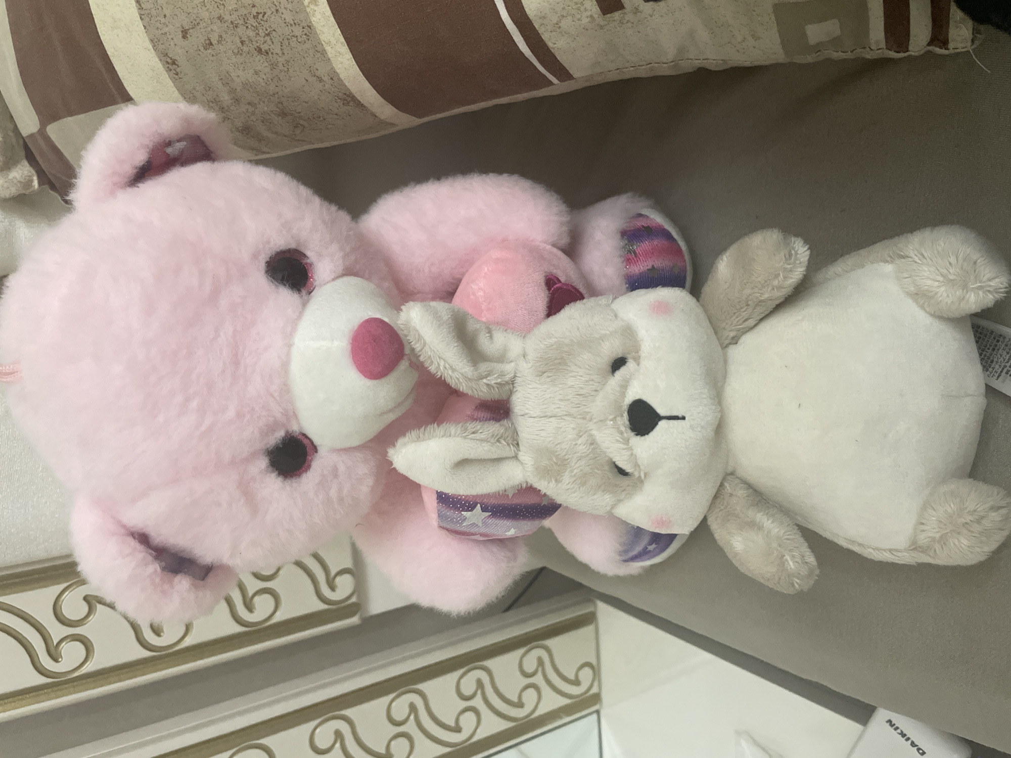 The scene depicts two plush toys positioned on a neutral-colored upholstered surface, likely a sofa, within an interior setting in the city of Ihtiman, Bulgaria. A larger, light pink teddy bear with dark, shimmering eyes, a white snout, and a pink nose is seated in the background. Its paws appear to be holding a smaller pink heart, and its feet feature a glittery, multi-colored striped pattern in shades of purple, pink, and white. In the foreground, a smaller, light gray and off-white bunny plush toy with dark eyes, a black nose, and a small black embroidered mouth sits directly in front of the bear. This bunny displays faint pink blush markings on its cheeks. To the left of the toys, a decorative white panel with gold-colored scrollwork is visible. Further to the left, a white object with the partially visible brand "DAIKIN" is present. To the right of the toys, a vertically striped pillow or cushion in tones of brown and beige is visible, contributing to the domestic environment. There are no individuals present.