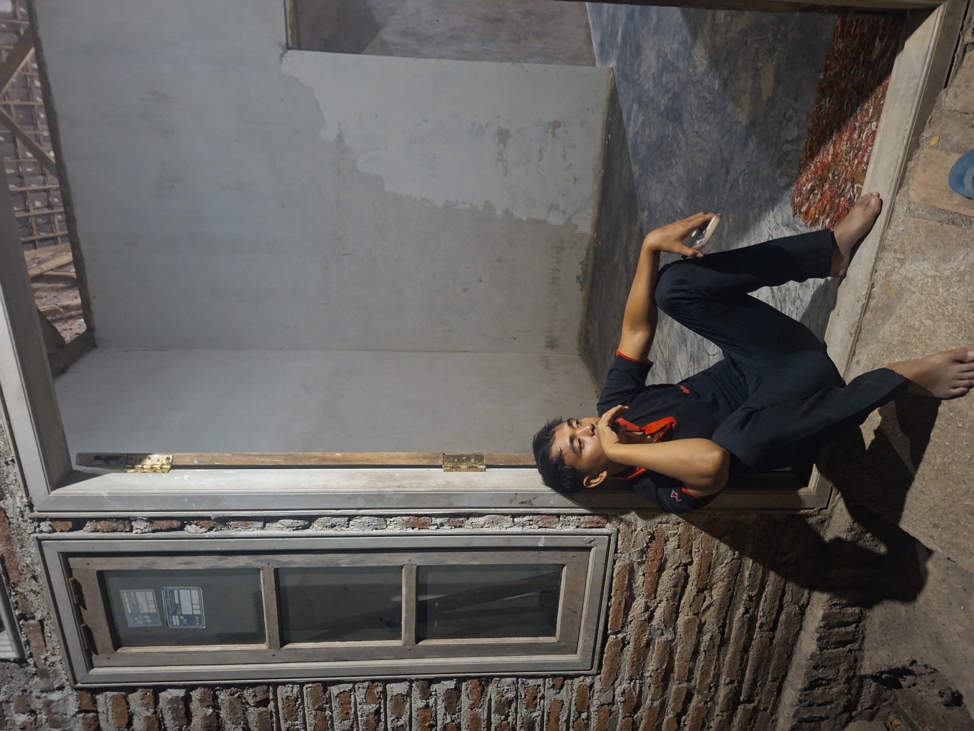 A man wearing a black polo shirt with red trim and black pants is sitting on a concrete step outside a building. He is leaning against a brick wall and looking to his right, with his chin resting on his hand. The building appears to be under construction, with exposed wooden beams visible in the ceiling. A window with wooden frames is set into the brick wall. The ground is a rough concrete surface with a patterned rug visible inside the doorway. The lighting suggests it is nighttime, with shadows cast by the man and the building. The location is identified as Tongaswetan, Indonesia.
