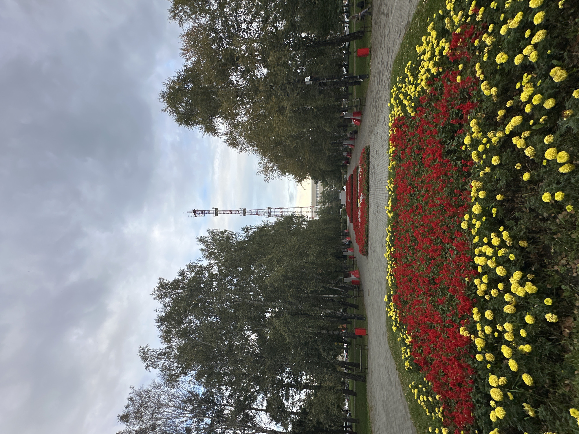 A wide-angle, eye-level shot captures a park in an unknown city under a cloudy sky. In the foreground, vibrant flowerbeds with yellow and red blooms are meticulously arranged. Beyond the flowers, a paved walkway stretches into the distance, flanked by mature trees on both sides. A tall, red and white radio tower stands prominently in the background, positioned centrally between the tree lines. Several park benches are visible along the walkway, with a few individuals seated on them. Red trash receptacles are also scattered throughout the park. The overall impression is one of a well-maintained urban green space.