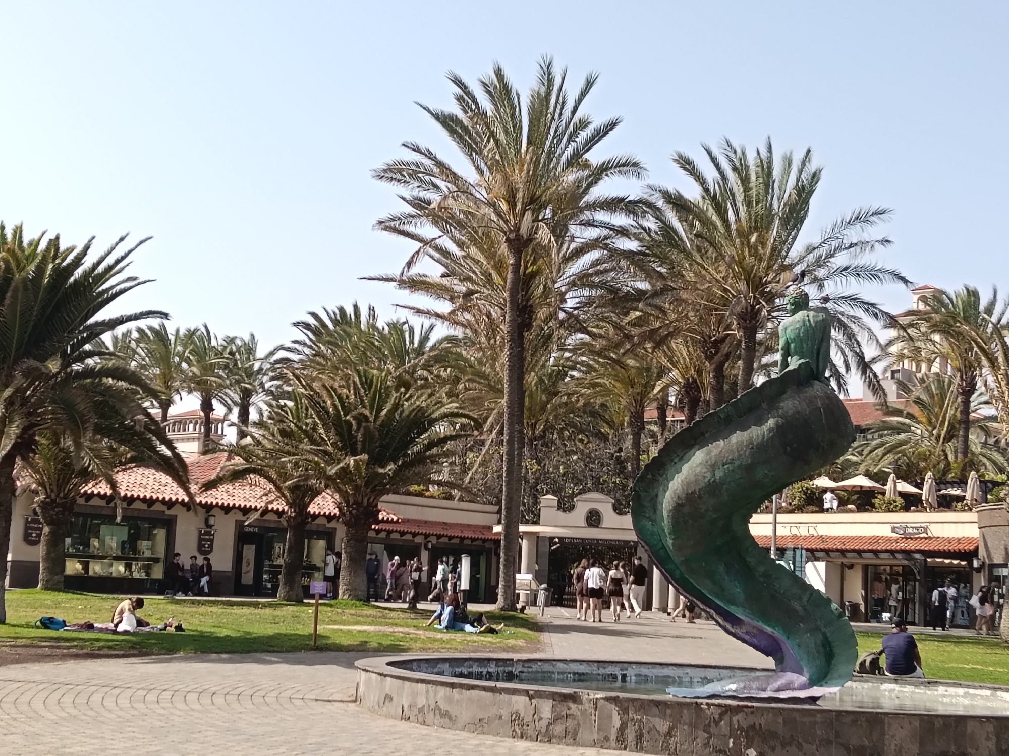 The image captures a sunny scene in Maspalomas, Spain. The foreground features a distinctive greenish-blue sculpture resembling a serpentine creature emerging from a low fountain. In the background, a building with a red-tiled roof hosts multiple storefronts and entrances. Palm trees of varying heights are scattered throughout the scene, lending a tropical ambience. People are seen in different activities: some are strolling along the walkways, others are lounging on the grass, and a few are sitting near the fountain. The overall scene suggests a relaxed, tourist-oriented environment.