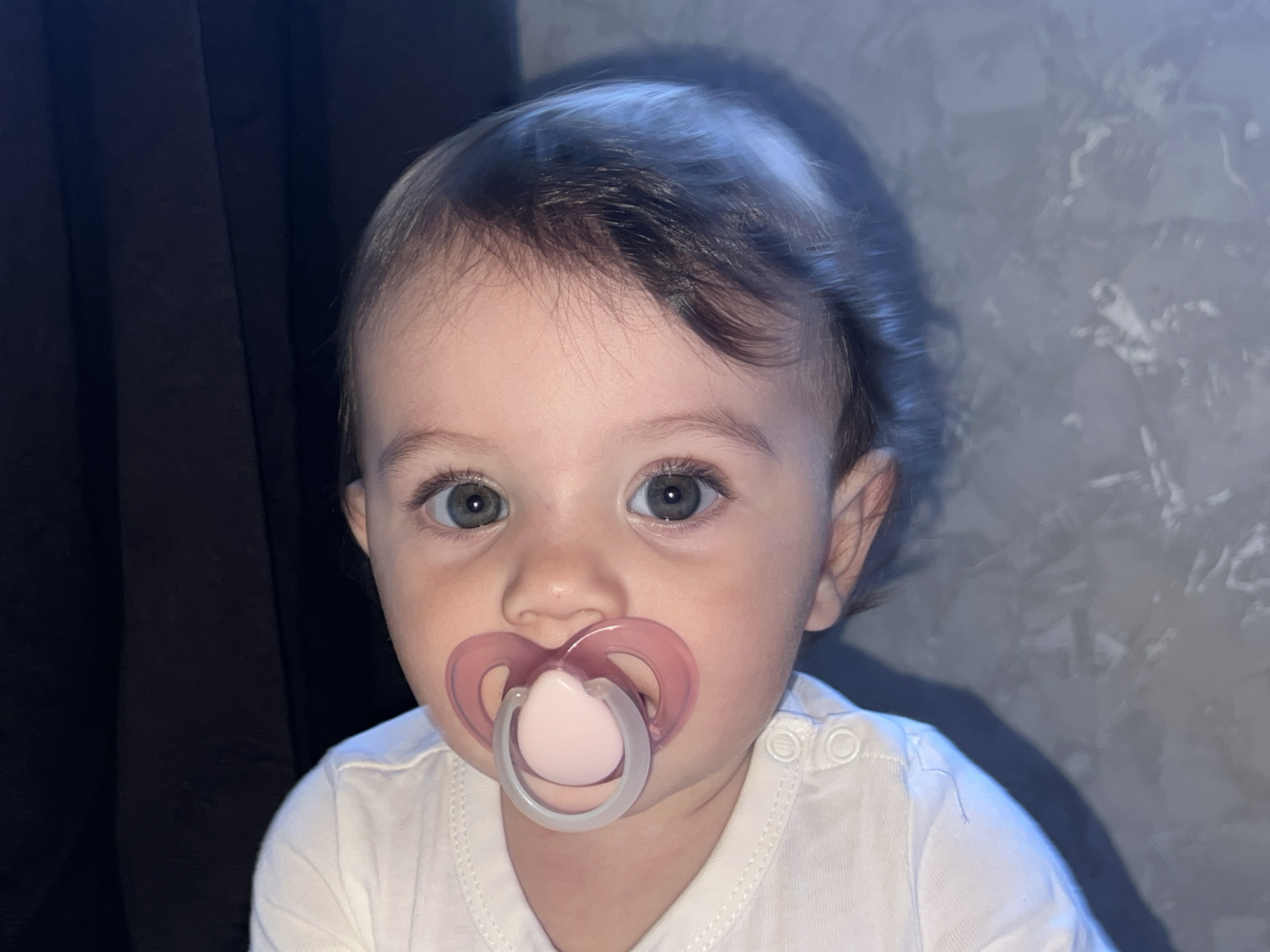 An indoor, frontal close-up depicts an infant, gender undetermined, with light complexion, dark hair, and blue-green eyes, looking directly forward. A pink and white pacifier is inserted into the infant's mouth. The infant wears a white upper garment featuring snap button closures on the right shoulder. The background consists of a dark, uniform fabric on the left and a light gray, textured wall on the right. A shadow cast from the infant's head and left shoulder is visible on the textured wall. The depicted scene is stated to be in the city of Elena, Bulgaria.