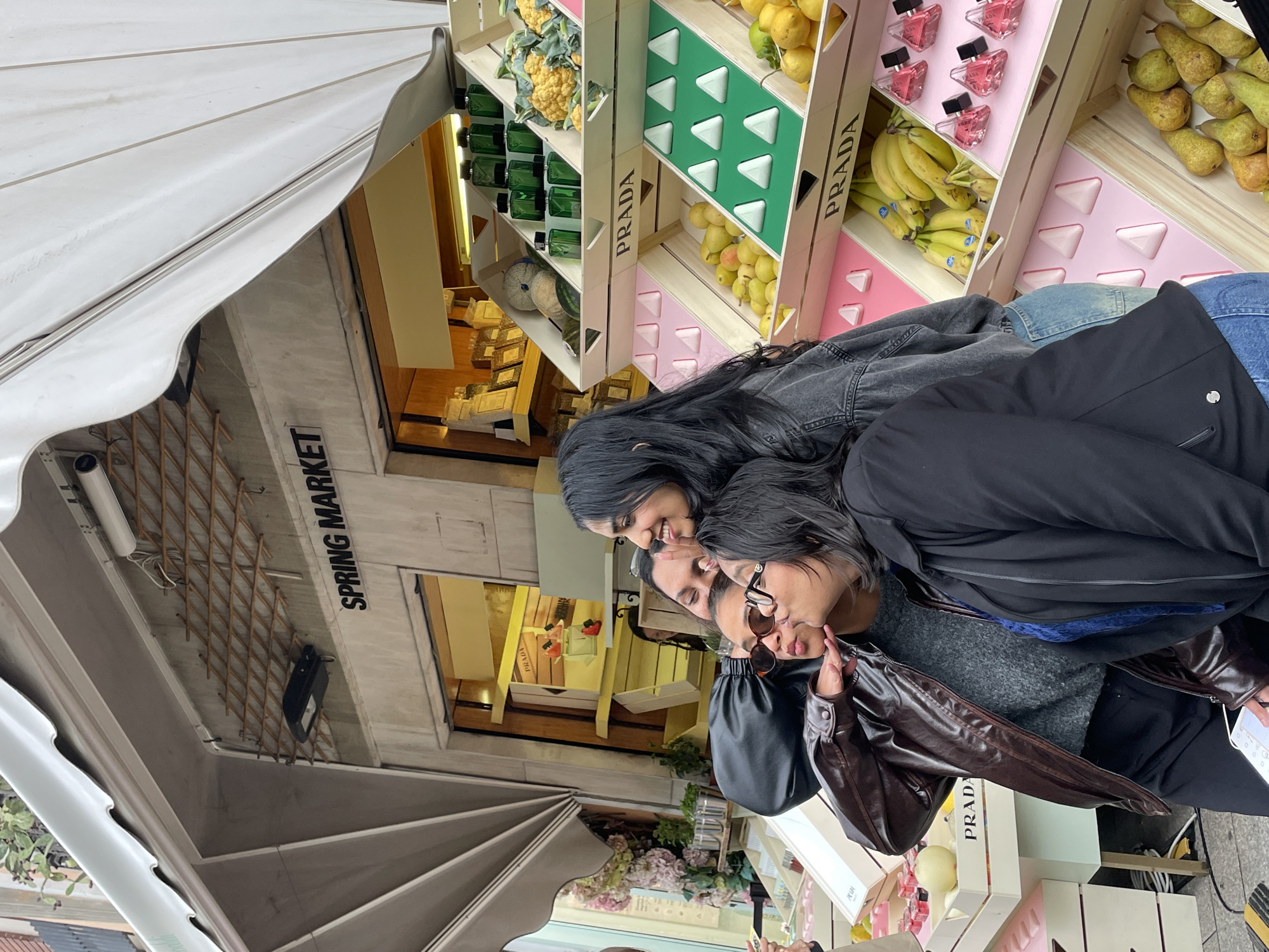 The image depicts two women posing in front of a shop display that includes stacked crates labeled "PRADA". The crates contain various objects including fruits, vegetables, and perfume bottles. The shop, labeled "SPRING MARKET", is located in Milan, Italy, evidenced by the architecture and shop name. The women appear to be taking a photo or selfie.