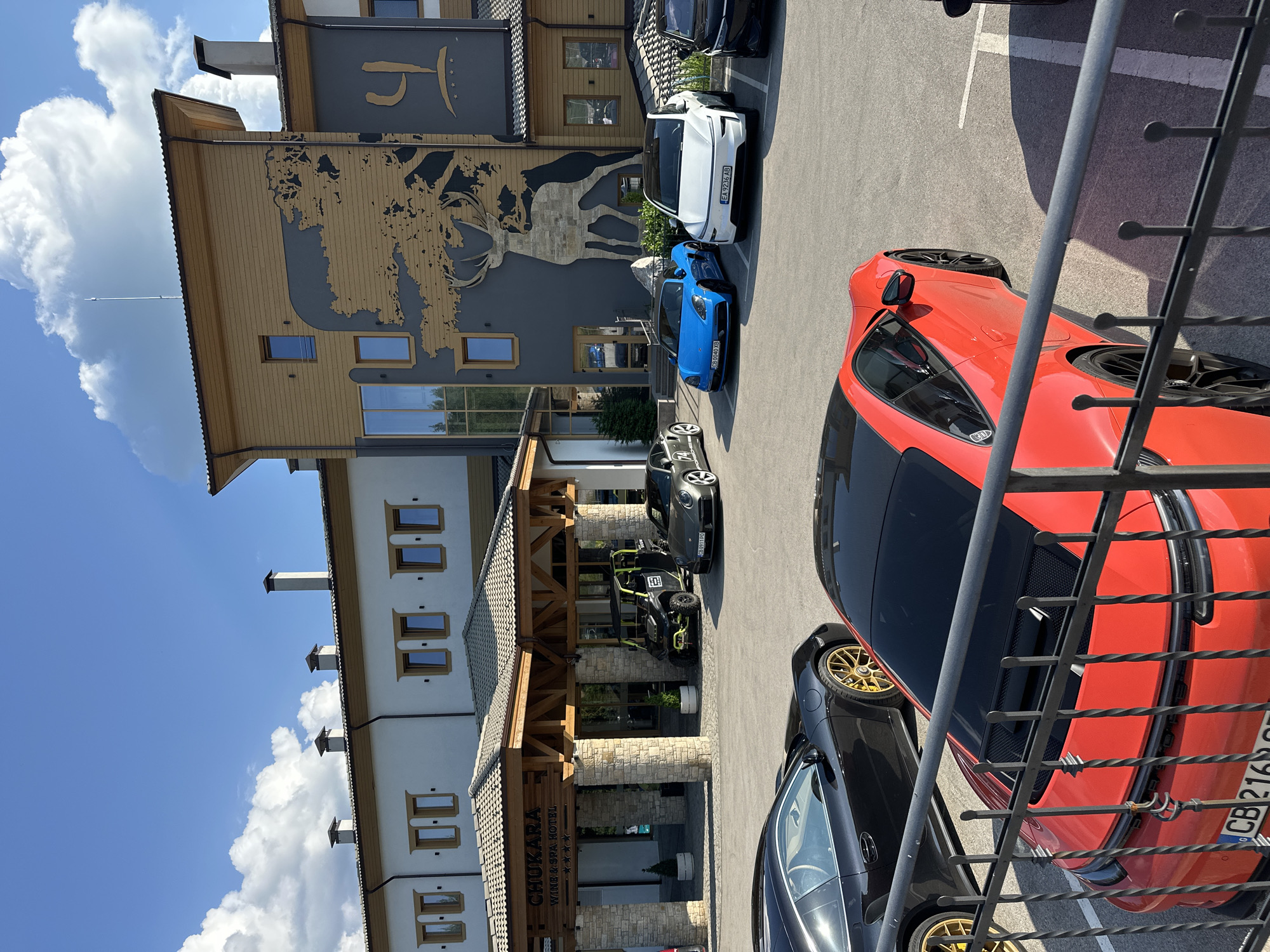 This is a medium shot taken from a slightly elevated angle, looking down at a hotel entrance and parking lot. The hotel, identified by the sign "CHUKARA WINE & SPA HOTEL", features a stone facade and a wooden-beamed entrance. Several cars are parked in the lot, including a bright red sports car with gold rims, a black sports car, a blue sports car, and a white Tesla. A lime green off-road vehicle is also parked near the entrance.
The building has a modern yet rustic architectural style, with multiple windows and chimneys visible. The background shows a bright blue sky with fluffy white clouds, suggesting a clear, sunny day, likely in the afternoon due to the shadows cast by the cars and railings. The surrounding environment appears to be a resort or hotel area, possibly in a mountainous region given the style of the building and the presence of a large mural of what appears to be a deer on the side of the hotel.
The overall impression is one of a luxurious getaway, with high-end vehicles hinting at the clientele. The image is oriented vertically, with the bottom of the frame showing the cars and the top showing the hotel's upper floors and the sky. The visible text includes the hotel's name and various license plate numbers such as "EA 9236 AB", "C80911P0", and "CB216".