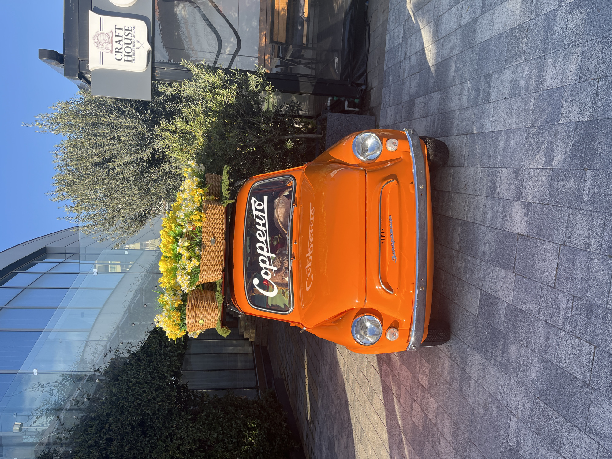 An eye-level, medium shot shows an orange vintage car parked on a paved surface. The car has flower-filled baskets attached to its roof. The name "Copperpen" is written vertically on the windshield. The front of the car faces the viewer. In the background, there is a multi-story glass building with a green vine climbing along its exterior wall. A sign for "CRAFT HOUSE THE BEST BEER & WINE" is visible above the car. The pavement is made of gray rectangular blocks, and sunlight creates shadows across the ground. The location is Sochi, Russia.