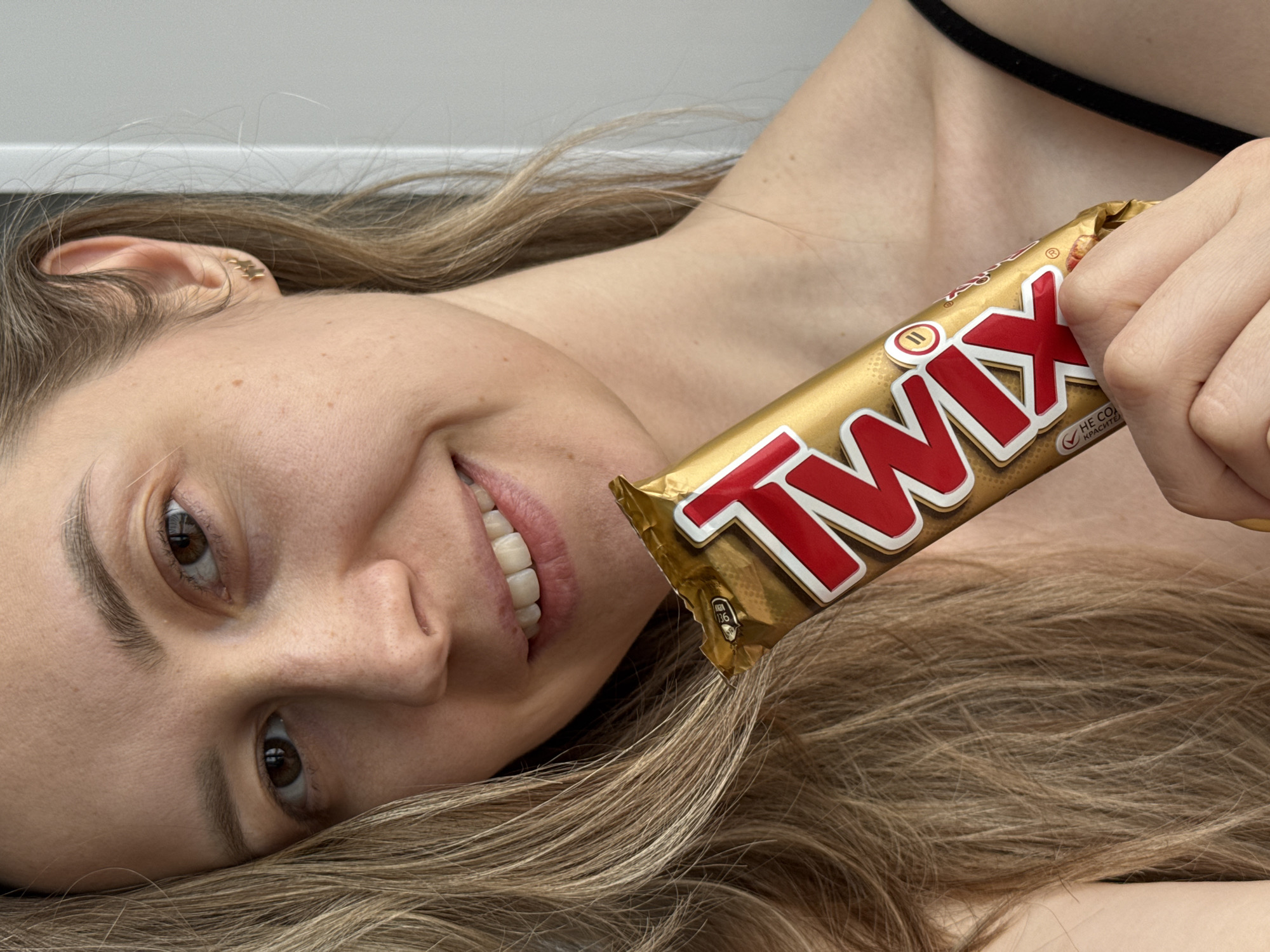 A woman lies on her back, looking up and smiling while holding a Twix candy bar. The location is not discernible from the image. The woman has fair skin, brown eyes, and light brown hair. She is wearing a thin black strap, possibly from clothing, on her upper chest. The candy bar is a gold-colored wrapper with the word "Twix" in red and white lettering.