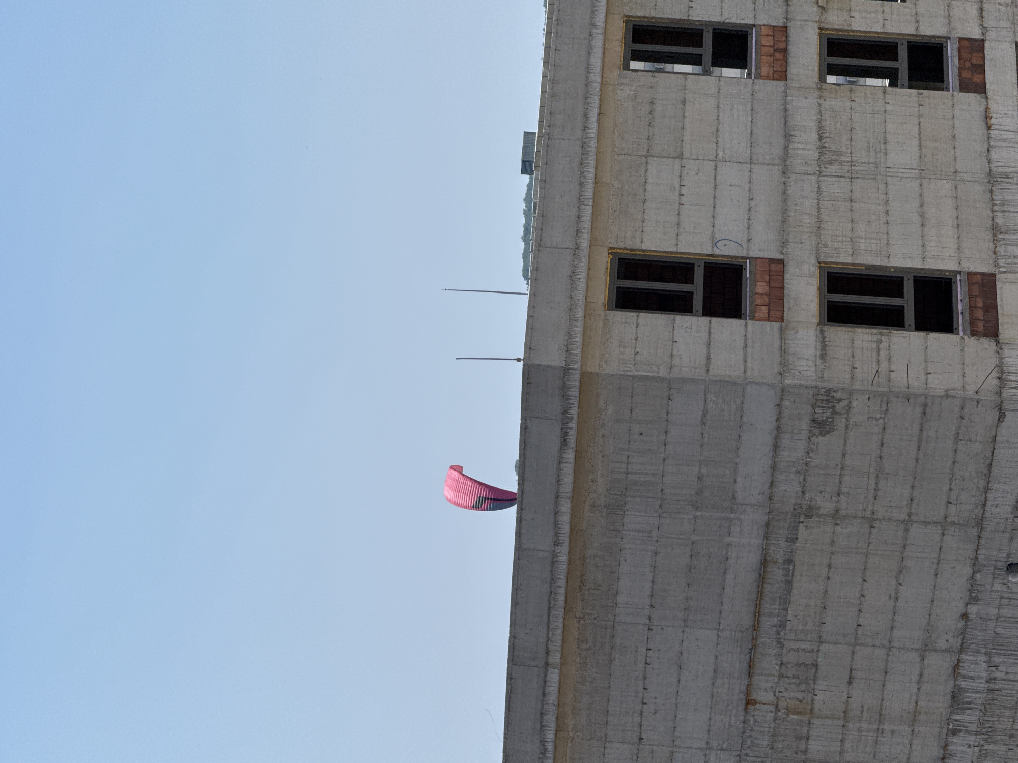 A pink and blue kite is airborne against a clear blue sky, positioned above a concrete building under construction in Boreti, Montenegro. The building features multiple windows, some with brick trim, and a rough concrete facade. The top of the building is flat and appears to be a roof. The kite is the primary focal point in the sky, while the building occupies the majority of the frame, emphasizing its unfinished state.