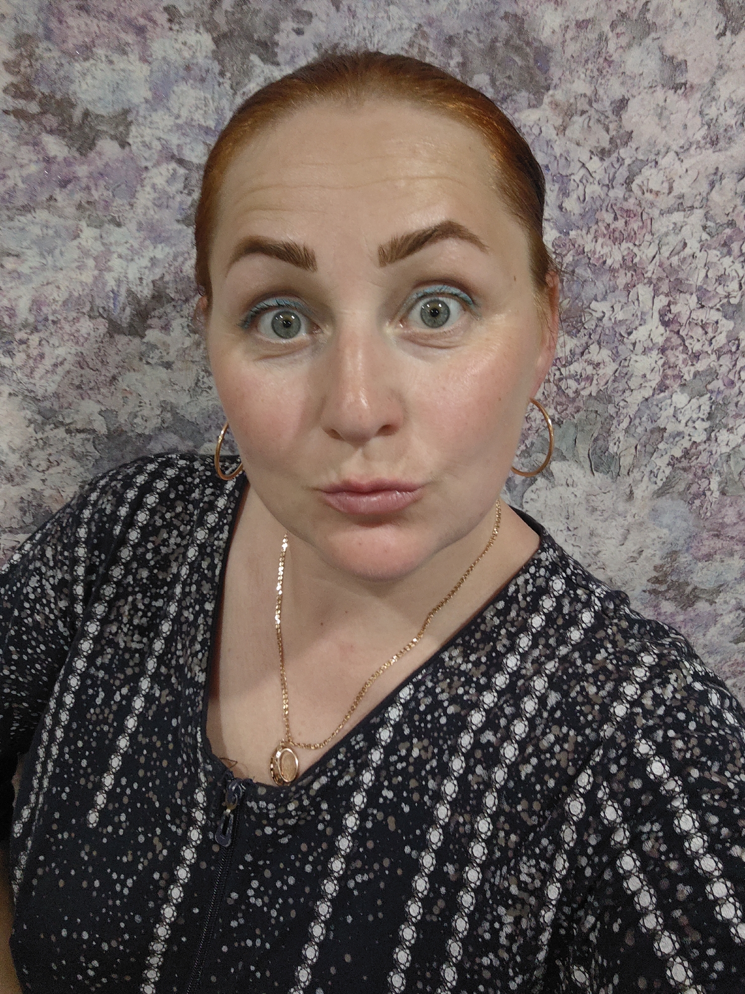 A head-and-shoulders selfie of a fair-skinned woman with reddish-brown hair pulled back and green eyes looking directly at the camera with a surprised expression and puckered lips. She is wearing a patterned black and white blouse with a visible zipper and gold jewelry including hoop earrings and a necklace with a pendant. The background is a textured wall with a mottled purple, gray, and white pattern. There is no specific information to suggest this photo was taken in Фоки, Russia.