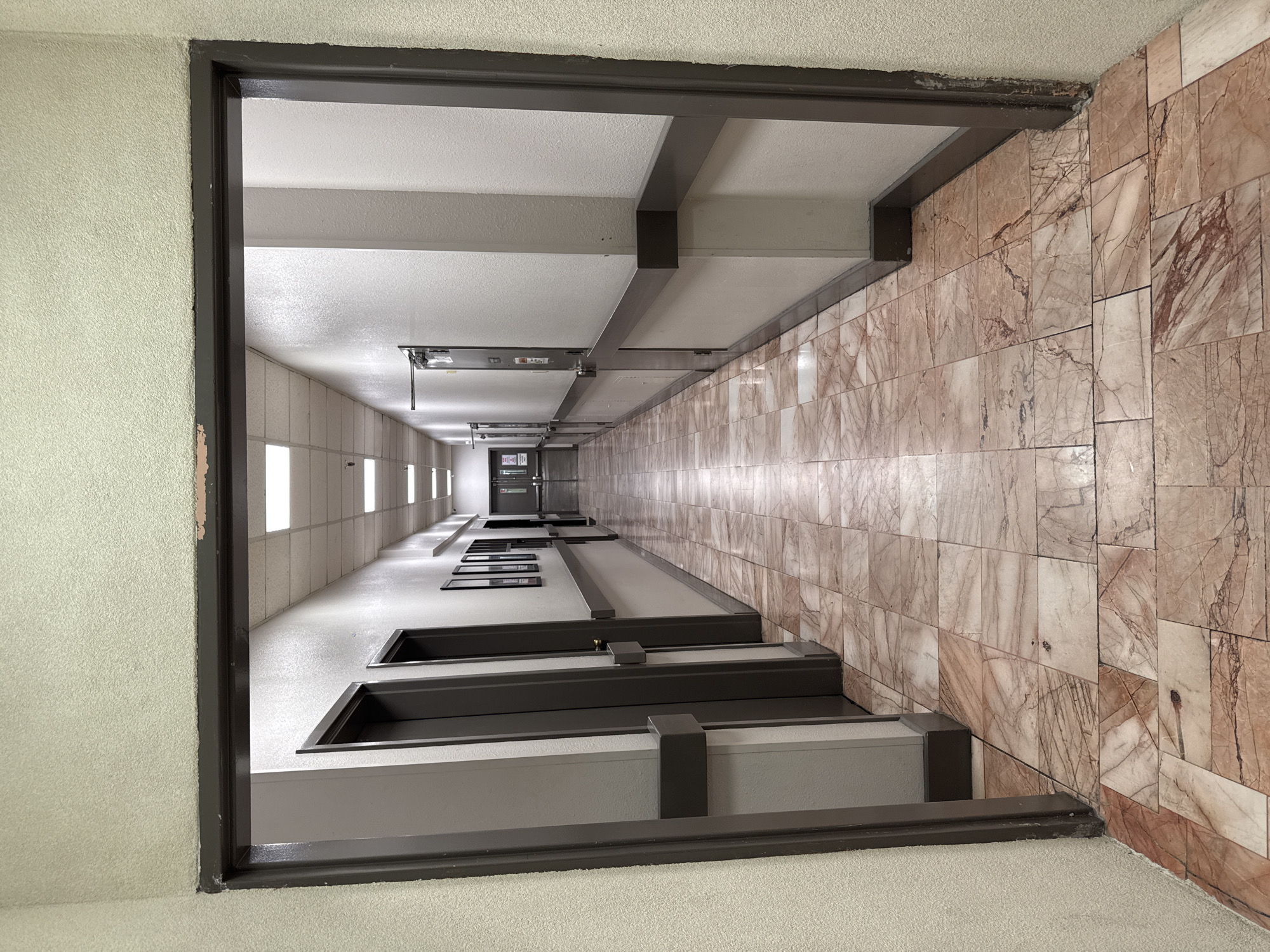 The image shows a long, empty hallway with a tiled floor. The hallway is viewed from an open doorway. The walls are painted a pale color, and there are doors on both sides of the hallway. The floor is tiled with a pattern of pinkish-brown and cream-colored tiles. There are fluorescent lights on the ceiling. The hallway extends into the distance, where it appears to turn a corner. The scene is well-lit and appears to be indoors. The location is in El Paso, United States of America.