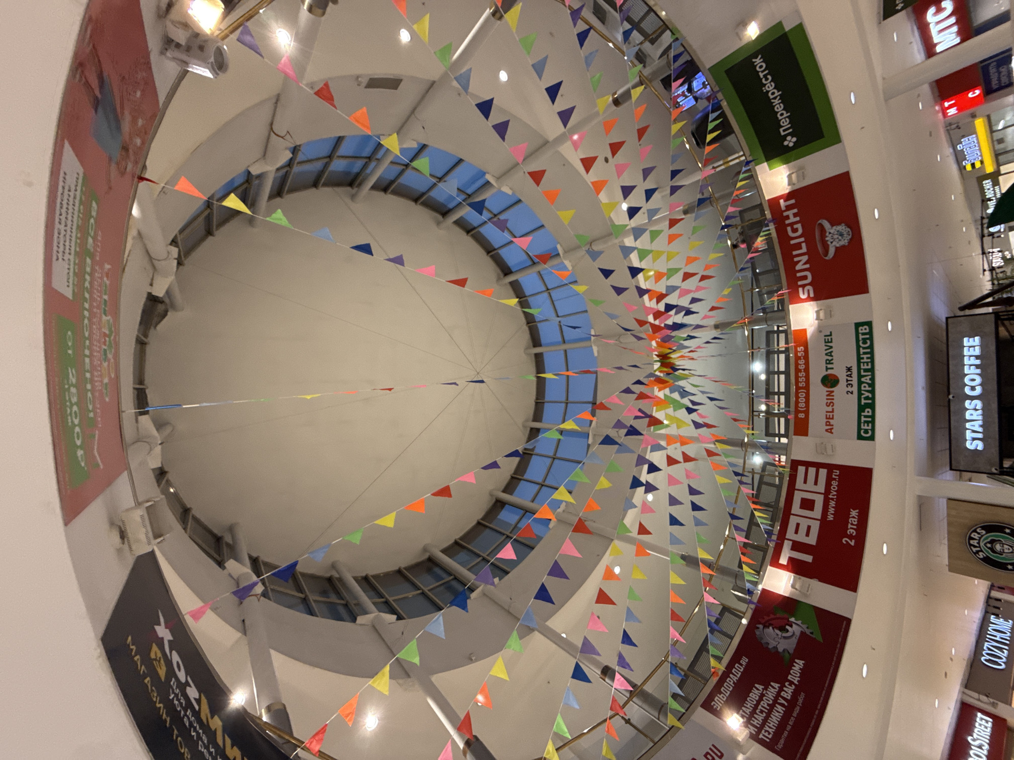The image shows the interior of a shopping mall in Moscow, Russia. The view is directed upwards towards a domed ceiling adorned with colorful triangular pennant flags. Below the ceiling, there are multiple levels of the mall visible, with various shop signs and storefronts lining the open walkways. The shop signs include "Перекрёсток", "SUNLIGHT", "TBOE", "STARS COFFEE", "COZY HOME", "Apelsin Travel", and "Эльдорадо.RU". The architectural design features a circular opening in the center, revealing a view of the sky through a grid of windows. The lighting is provided by ceiling lamps and natural light from the window grid.