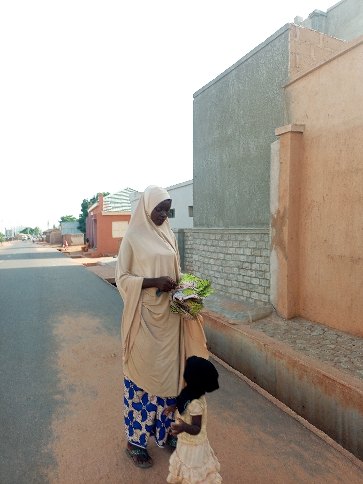 A woman and child are walking on the road in Tempure, Nigeria. The woman is wearing a tan hijab and gown, and blue patterned pants. The child is wearing a light-colored dress and head covering. The woman is carrying a bundle of green fabric. They are on a road next to buildings. The road is paved, and there is a shallow ditch between the road and the buildings. One building is unfinished, with exposed concrete. Another building is painted reddish-brown. Other buildings and trees are visible in the distance. The sky is bright.