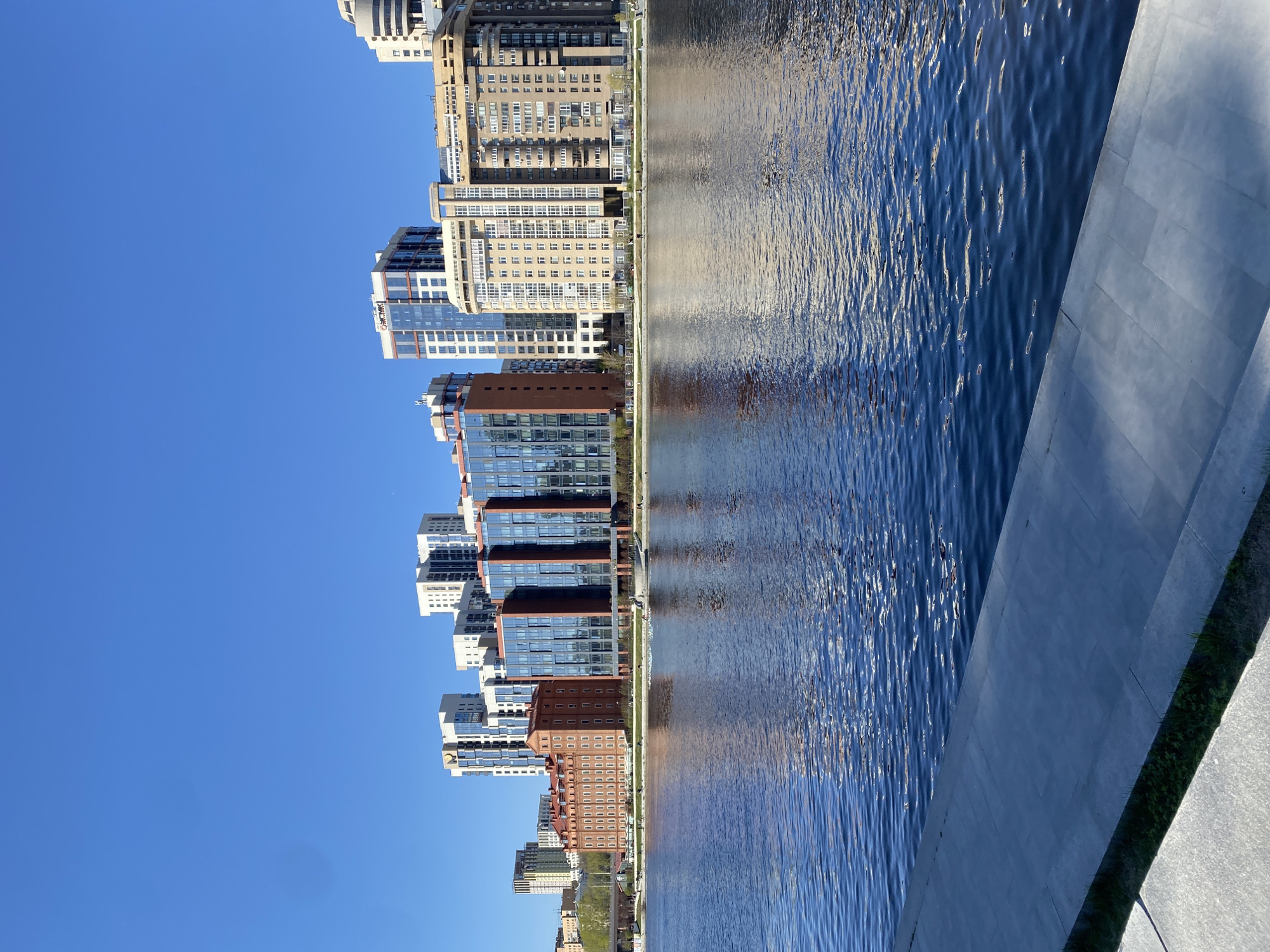 A vertically oriented photograph depicts a waterfront scene in Yekaterinburg, Russia. The left side shows a series of modern, high-rise buildings of varying architectural styles, including structures with glass facades and brick exteriors. The buildings are reflected in the water on the right side of the image. The sky is clear and blue. A concrete embankment and hints of green vegetation are visible at the bottom right corner.