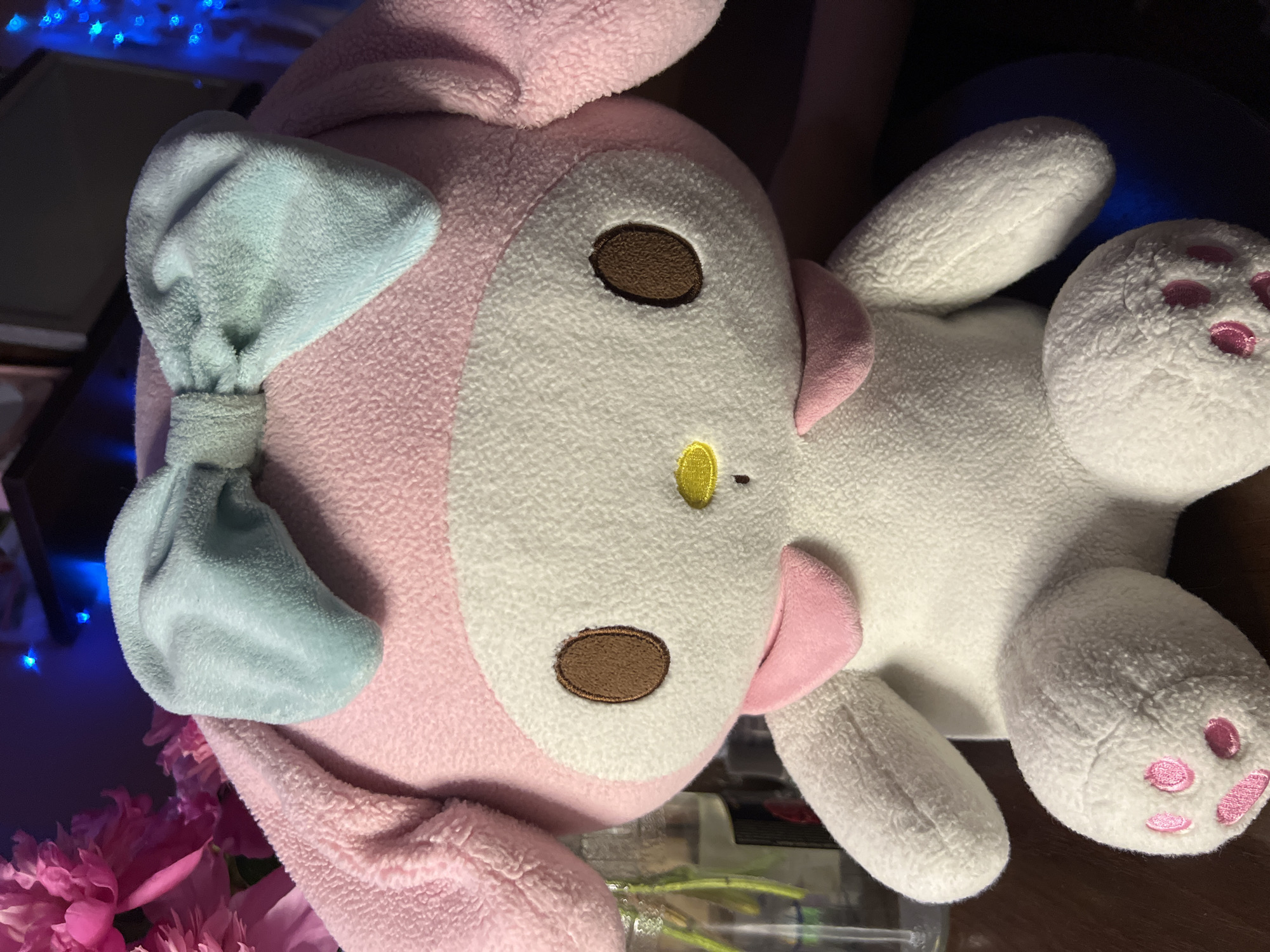 A close-up, eye-level, medium shot captures a plush toy, likely My Melody from Sanrio, positioned centrally. The toy is predominantly pink and white with a light blue bow atop its head. Its features include large, oval brown eyes with darker brown stitching, a small yellow nose, and a pink collar. The background is dark and appears to be indoors, with hints of pink flowers on the left and blue lights on the right, suggesting decorative elements or ambient lighting. The toy is resting on a dark wooden surface. The image is sharp and well-lit, focusing on the toy's textures and details. There is no discernible connection to Minsk, Belarus in the visual information provided.