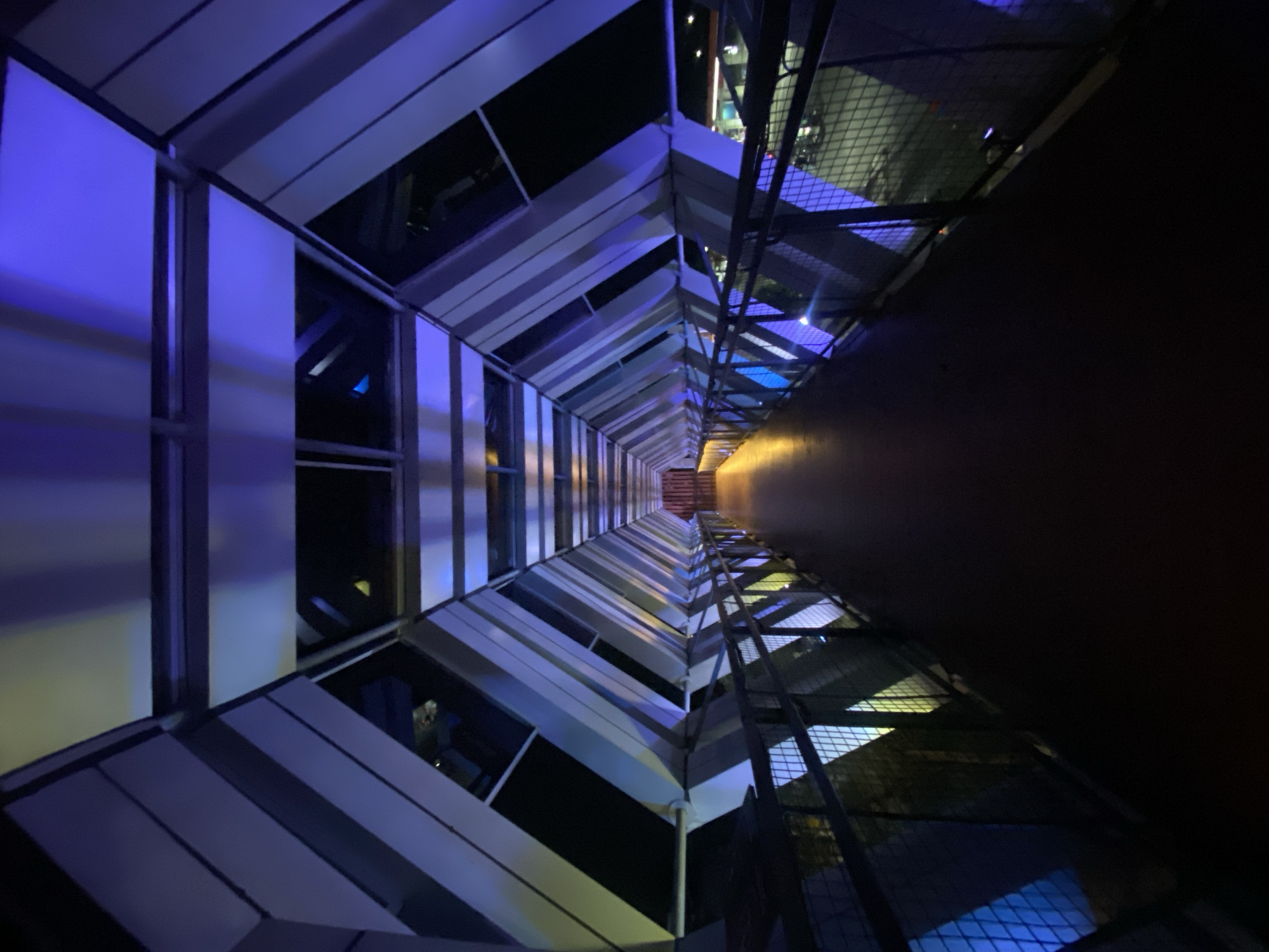 The image shows a view from inside a pedestrian bridge, looking down a long, enclosed walkway. The bridge has a futuristic design with angled panels and illuminated sections. The walkway is dark, with the floor appearing to be a smooth, dark surface. The sides of the bridge are lined with metal railings and mesh panels, through which some outside lights and structures are visible. The ceiling of the bridge is made up of a series of angled panels, some of which are illuminated with a vibrant blue and purple light, creating dramatic shadows and reflections. The perspective draws the viewer's eye down the length of the bridge towards a dimly lit opening at the far end. The overall impression is one of modern architecture and atmospheric lighting, creating a tunnel-like effect.
This specific pedestrian bridge is located in Banjarbaru, Indonesia, connecting the Syamsudin Noor Airport to the nearby hotel.