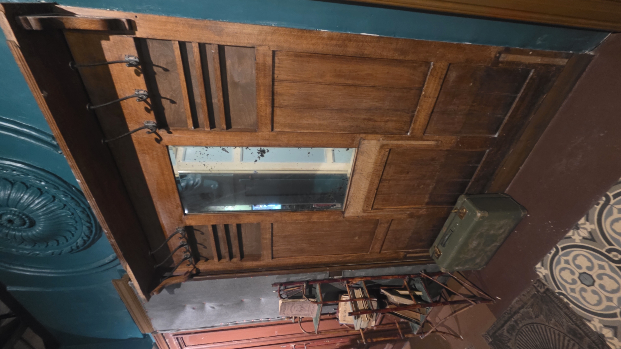 The image shows an interior scene, appearing to be the ceiling of a room. The ceiling is decorated with an elaborate wooden structure with a mirror inset, suggesting a hat and coat rack. There are metal hooks attached to the structure, presumably for hanging clothes. Below the ceiling fixture, there is a small metal rack holding books or papers. A green suitcase rests on a surface adjacent to the wooden structure. The environment is detailed with patterned tiling on the floor and decorative elements on the ceiling, indicating an older or historic building. The scene appears to be in Saint Petersburg, Russia, based on the architectural style and details typical of buildings in that city. The scene is inanimate, with no people present.
