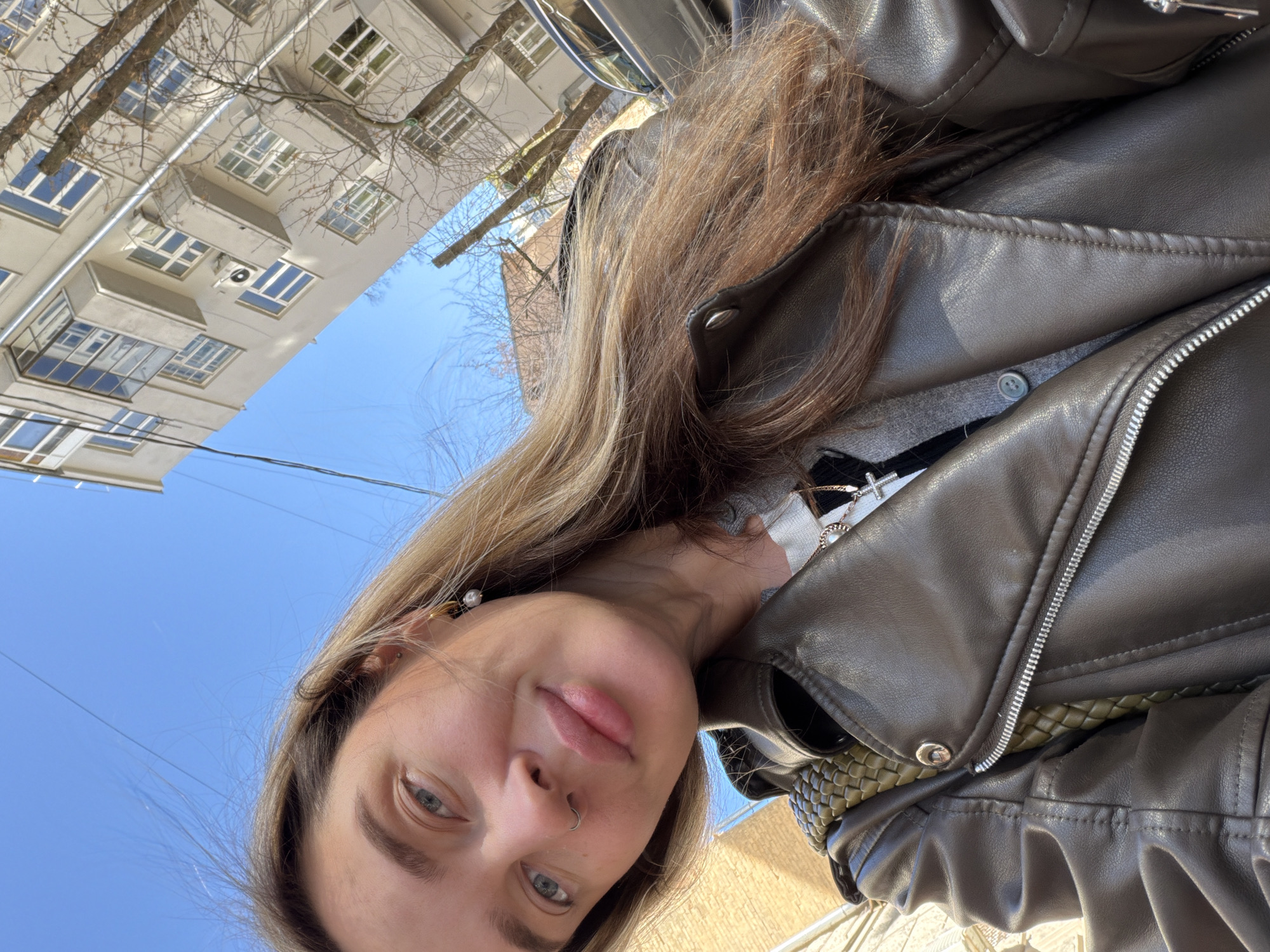 A woman with fair skin and light brown hair is seen in a close-up selfie, her face angled toward the camera, which is tilted upward to include a blue sky and a building in the background. She has blue eyes and a piercing in her left nostril. She wears a dark brown leather jacket over a grey sweater and has a pearl earring on her left ear. The building behind her is cream-colored with multiple windows and tree branches visible in front of it. The picture appears to be taken outdoors, likely during the day. Based on architectural style and context, the location is consistent with urban areas in Moscow, Russia, though more specific identifiers are absent in the image.