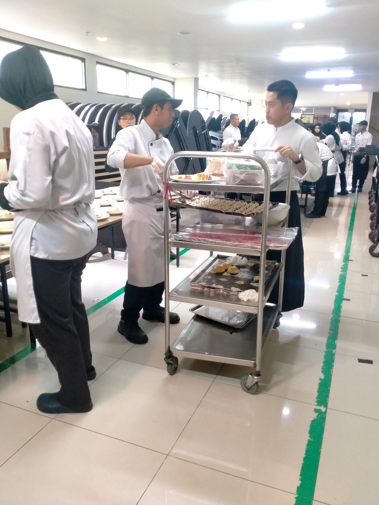 In a brightly lit indoor setting, possibly a culinary school or restaurant kitchen in Batu, Indonesia, several individuals are engaged in food preparation and service. Two men in white chef uniforms and aprons are the primary focus. One, on the left, wears a green cap and appears to be arranging food on a cart. The other, on the right, is also in a white chef's uniform and is seen pushing a multi-tiered stainless steel cart laden with trays of food, including what appear to be pastries or desserts. A person wearing a dark hijab and white uniform is visible in the foreground to the left. Other individuals in chef attire are present in the background, suggesting a group activity. The environment features tiled flooring with a prominent green line, windows, and stacks of chairs. The overall scene depicts a busy, organized food service operation.