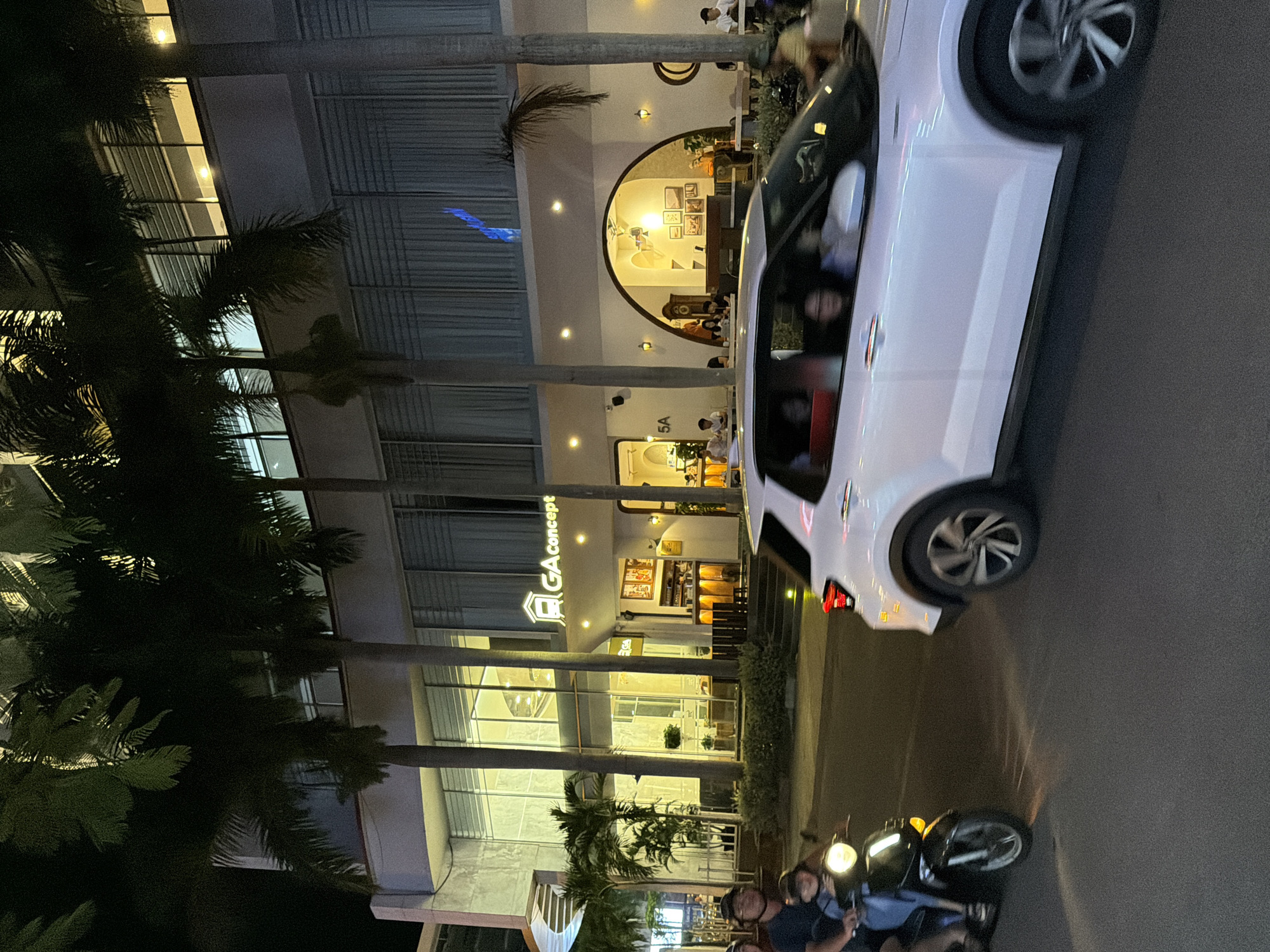 The image captures a nighttime street scene in Nha Trang, Vietnam, showcasing the facade of a modern building with a prominent "GA concept" sign. Palm trees line the street, their fronds silhouetted against the illuminated building. A white SUV is seen in motion on the right side of the frame, its wheels blurred due to the speed. On the left, a black scooter carries two people. The building itself features large glass windows and an arched entrance leading to a well-lit interior with tables and chairs, suggesting a cafe or restaurant. The overall ambiance is urban and commercial, illuminated by artificial lighting.
