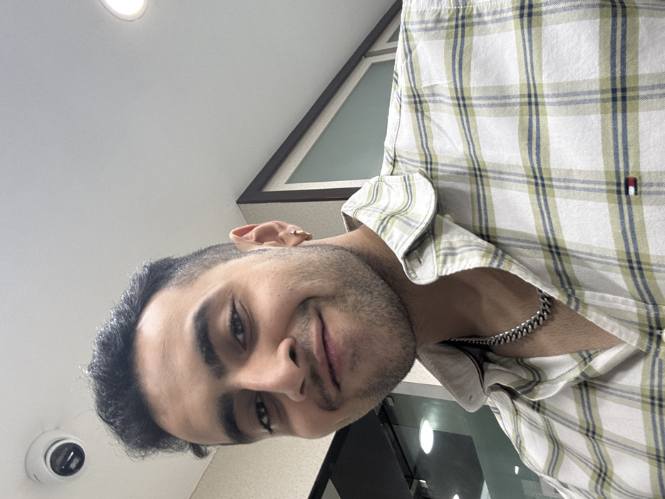 A man smiles subtly at the camera. He has dark hair, brown eyes, and stubble. He is wearing a white and light green plaid shirt. A silver chain necklace is visible around his neck. In the background, there is a window with a dark frame and a white ceiling with a security camera in the corner.