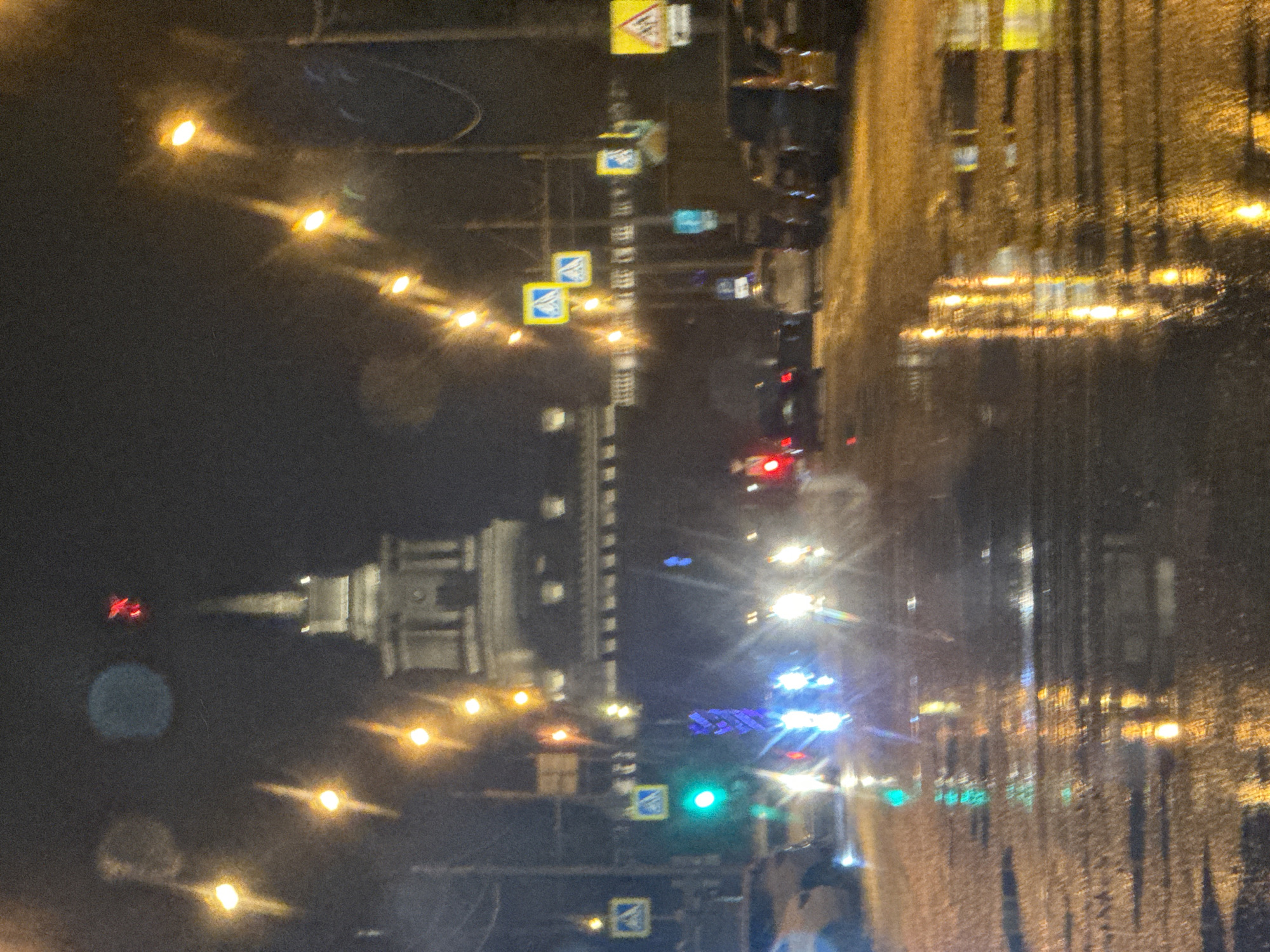 The image depicts a nighttime view of Krasnoyarsk, Russia, captured through a wet window. The focus is on the city skyline, featuring the Saint Pokrovsky Cathedral, illuminated streetlights, and traffic signals. Cars are visible on the road, indicated by their headlights and taillights. Street signs are also visible. The overall environment suggests a rainy or wet urban setting at night.