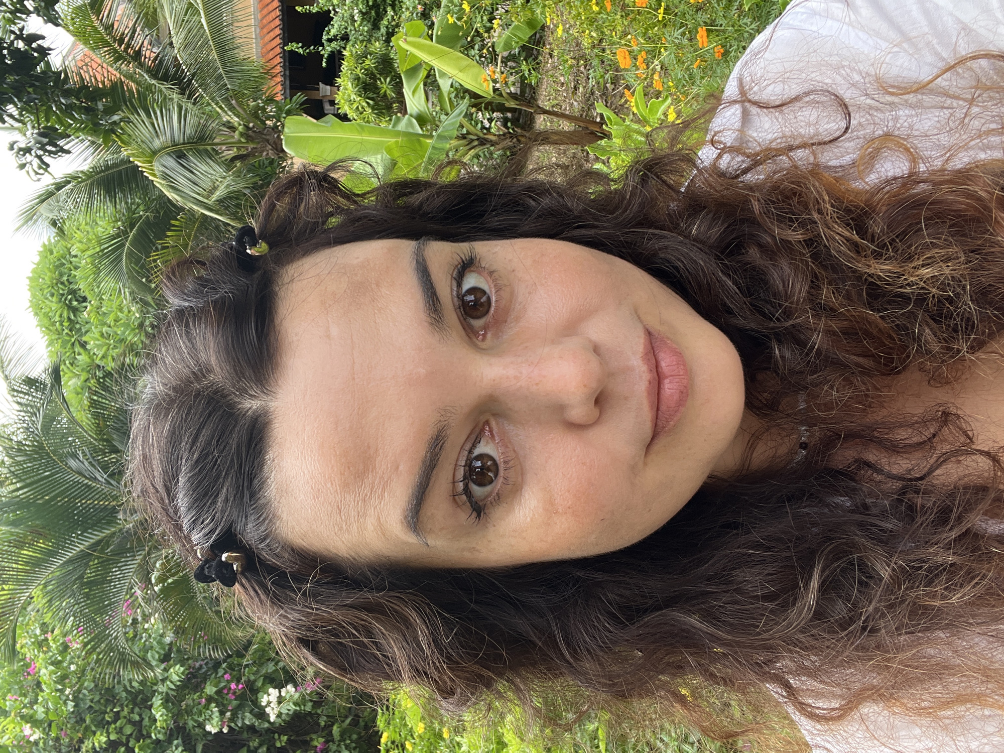 A medium close-up selfie features a woman with long, curly brown hair. She is wearing a white shirt, and her hair is partially held back on either side of her head by small, black butterfly-shaped clips with gold accents. The woman has brown eyes, fair skin with some freckles, and light pink lips. She is looking directly at the camera.
The background is lush and green, suggesting an outdoor, tropical environment. There are palm trees with large fronds, as well as smaller trees and flowering plants with yellow and pink blossoms. In the distance, a building with an orange-tiled roof is visible. The overall impression is one of a warm, humid climate, consistent with a location in Phu Thuy Ward, Vietnam, known for its tropical atmosphere and natural beauty.
