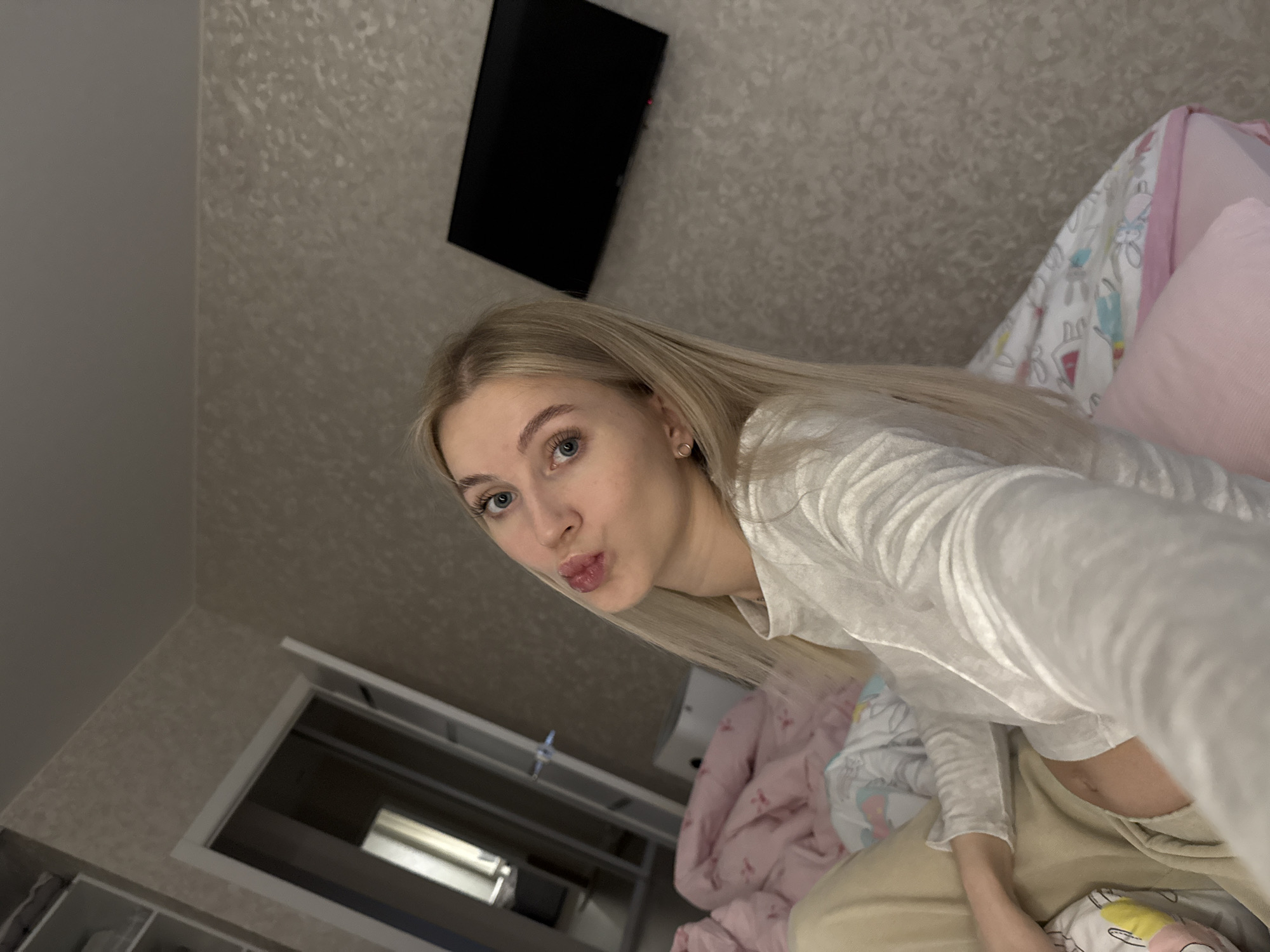 A selfie taken indoors shows a fair-skinned young woman with blonde hair and blue eyes making a kissing face. She is wearing a long-sleeved white shirt and is situated on a bed with pink pillows and a patterned blanket. In the background, there is a black television mounted on the wall and a white shelving unit with open compartments. There is no indication of location.