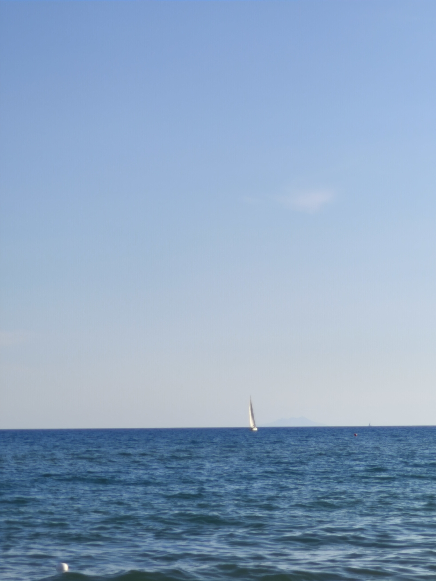 The media file depicts a serene seascape with a clear, pale blue sky dominating the upper portion of the image. A vast expanse of deep blue ocean fills the lower half, with gentle waves creating a textured surface. On the horizon, a small white sailboat with its sail fully extended is visible, appearing to be in motion. Further in the distance, another, smaller sailboat can be seen, and a red buoy marks a point in the water. A faint outline of a distant landmass, possibly an island, is visible on the horizon behind the sailboat. In the foreground, at the very bottom of the image, a white object, likely a buoy or part of a marker, partially submerged in the water, is visible. The lighting suggests it is daytime, likely morning or late afternoon, given the soft quality of the light and the absence of harsh shadows. The overall impression is one of tranquility and peacefulness. The location context provided indicates this scene is Castiglione della Pescaia, Italy. There is no visible text in the image.