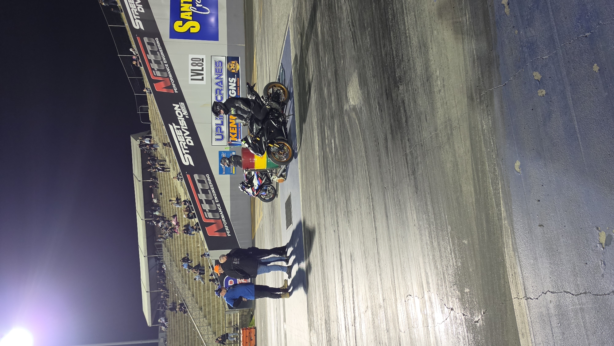 At night, three men stand on a drag strip in Eastern Creek, Australia, near two motorcycles. The men are dressed casually, with two in jackets and one in a blue long-sleeved shirt. The motorcycles are a black sportbike with a rider in full gear, and a white and blue sportbike parked behind it. In the background, spectators are seated in bleachers, illuminated by bright stadium lights. Various sponsor banners, including "Nitto Performance Engineering" and "Street Division by Nitto", are visible. The drag strip is paved and shows signs of wear, with tire marks and cracks.
