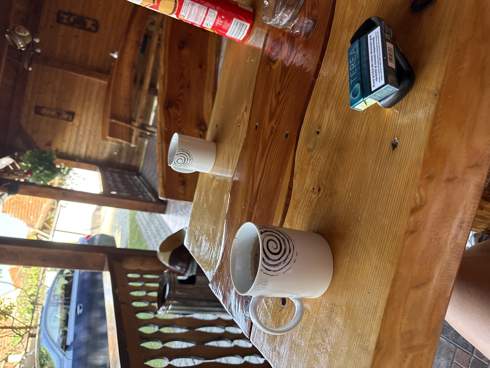 A wooden table on a covered patio holds two white mugs, one with dark liquid, a red bag of snacks, an ashtray with a cigarette, and a teal-colored pack of Terea. A blue car is parked on a paved driveway in the background, with a wooden railing and more wooden structures visible. The scene appears to be outdoors in Cârțișoara, Romania, on a sunny day.