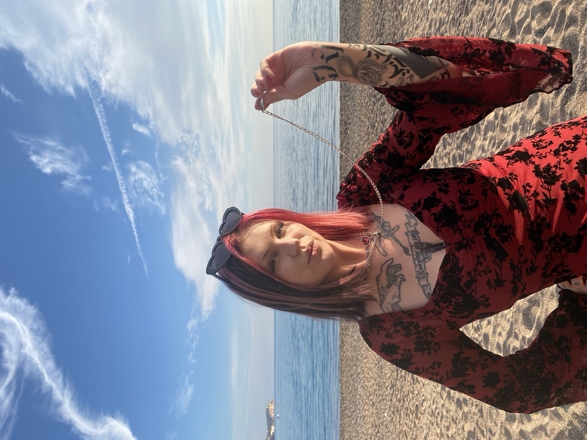 A woman is depicted standing on a sandy beach in Rafailovići, Montenegro. She is centered in the frame, directly facing the viewer with a slight pout. Her hair is dark brown with prominent pink streaks, and black heart-shaped sunglasses are perched on top of her head. She wears a long-sleeved red dress with a black floral pattern. Numerous tattoos are visible: a depiction of hands on her upper chest, script reading "Everything you can" below it, and extensive designs covering both arms, including an octopus-like figure and a lotus flower on her right arm. A silver chain necklace with a red heart-shaped pendant is around her neck. Her right hand is raised, holding a separate thin silver chain, while her left hand rests on her hip. The background features the calm, blue Adriatic Sea with a distant landmass visible on the left horizon under a clear blue sky dotted with scattered white cirrus clouds and visible contrails. The foreground consists of light brown sand interspersed with small pebbles.