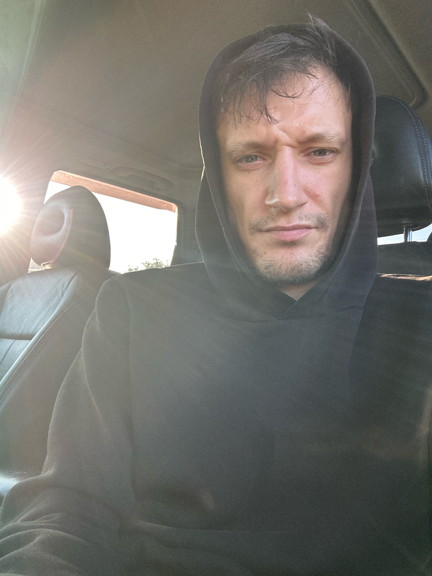 A male, appearing to be in his late 20s or early 30s, is captured in a close-up, selfie-style shot from inside a vehicle. He is wearing a dark, hooded sweatshirt with the hood pulled up, obscuring part of his hair and framing his face. The man has a stern expression, with a furrowed brow and direct eye contact with the camera. His facial features include prominent cheekbones, a slightly unkempt beard, and tired-looking eyes.
The setting appears to be daytime, with bright sunlight streaming in from the left side of the frame, casting lens flare and illuminating the interior of the car. The background shows a glimpse of car seats and a window, through which trees and a bright sky are visible. The sun's rays are also reflecting off the car's interior surfaces. The context of Makiv, Ukraine, is not visually discernible from the image itself. The overall impression is one of a candid moment, perhaps during a drive, with the harsh sunlight creating a dramatic effect.