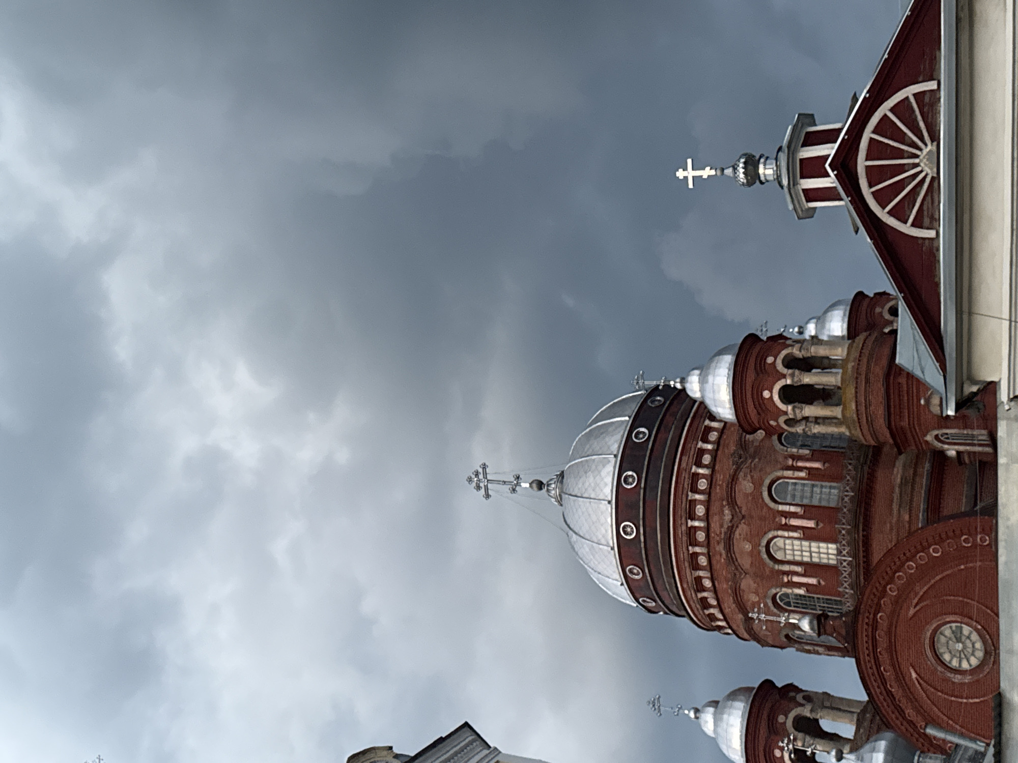 The image depicts a close-up, low-angle view of the domes and upper portions of the Trinity Cathedral in Urzhum, Russia. The cathedral is constructed of red brick with silver domes, each topped with a cross. The sky is heavily clouded with dark, dramatic storm clouds, suggesting overcast or impending inclement weather. The architecture features multiple smaller domes surrounding a larger central dome, with arched windows visible on the main structure. The detailed stonework and decorative elements of the cathedral are clearly visible against the dark sky.
