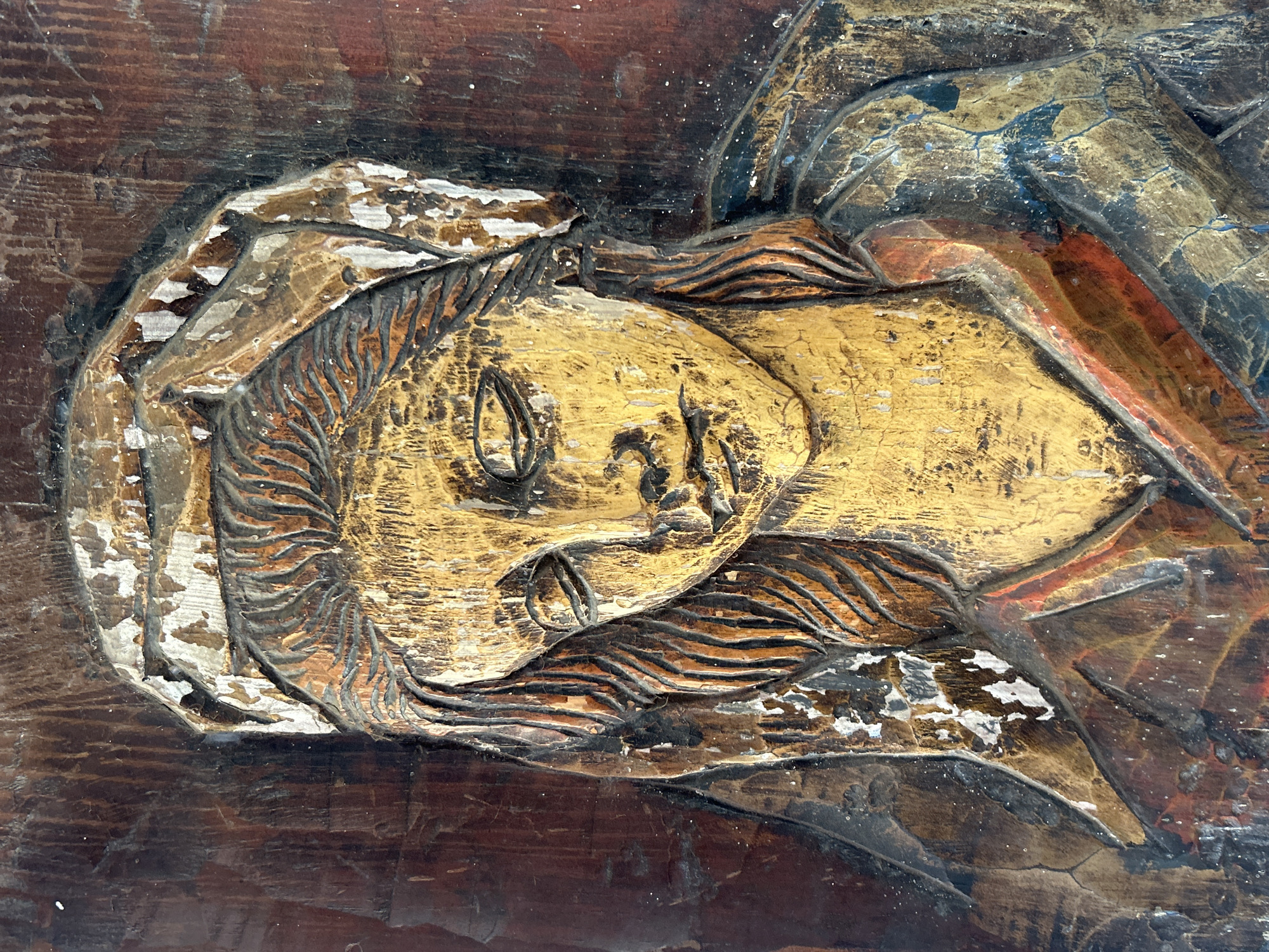 The image is a close-up, low-angle, medium shot of a distressed wooden carving of a woman's face. The carving, likely part of a larger sculpture or architectural element, is weathered and shows significant wear, with peeling paint and visible wood grain. The woman has her eyes closed, her head tilted downwards, and her expression appears somber or peaceful. She wears a headdress that is also chipped and worn, revealing the wood beneath. The carving is rendered in earthy tones, predominantly browns and yellows, with hints of red and blue in her clothing. The background is a dark, textured wood. This particular carving is located on the facade of the Cathedral of Our Lady of Carmen in Santa Ana, Venezuela.