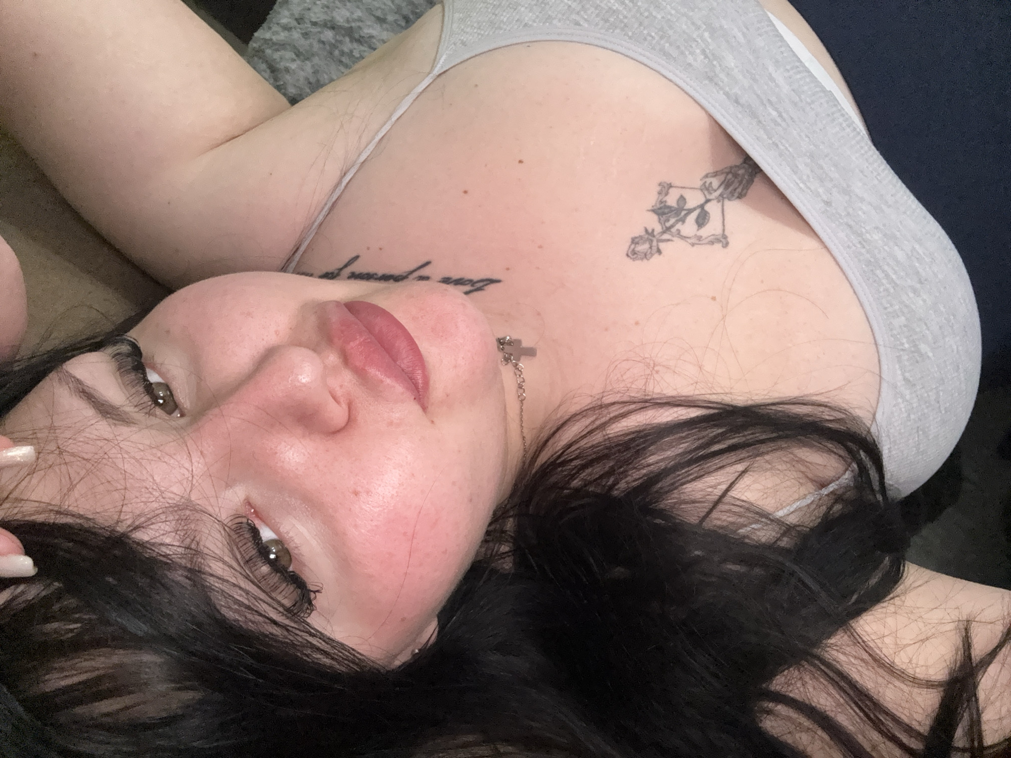 A woman with fair skin and dark hair is lying down, facing upwards. She has dark eyebrows and eyelashes, and light brown eyes. Her lips are full and pink. She has a tattoo of a skeleton hand holding a rose on her upper right chest, and another tattoo on her upper left chest. She is wearing a grey tank top. The background is dark and out of focus. The location is not discernible from the image.