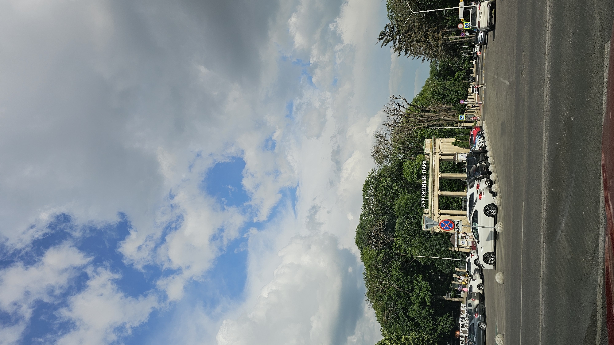 This image depicts a view of the entrance to Kurortny Park in Yessentuki, Russia, as seen from a street. The entrance is a large, ornate structure with columns and an archway, topped with the words "КУРОРТНЫЙ ПАРК" (Resort Park) in large lettering. In front of the park entrance, there is a parking lot filled with various cars. Several people are visible walking and standing near the entrance and in the parking lot. The scene is set against a backdrop of lush green trees and a sky with a mix of blue patches and white and gray clouds. The street in the foreground shows traffic lanes and a partial view of a vehicle. Traffic signs and streetlights are also present.