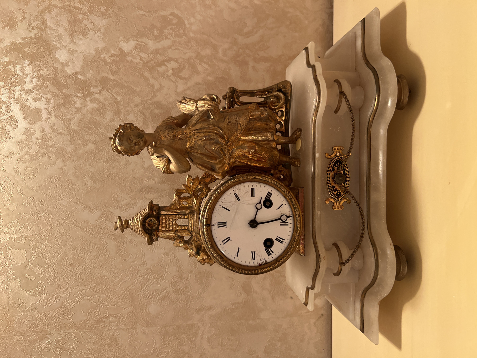 The image shows an ornate antique mantel clock with a golden figurine of a young girl holding a bundle of wheat. The clock itself is gilded and features Roman numerals on its white face. It rests on a white marble base with decorative gold accents. The background is a textured, light-colored wall. The clock is positioned on a light-colored surface, likely a table or mantelpiece. There is no indication of the city of Bender, Moldova in the image.