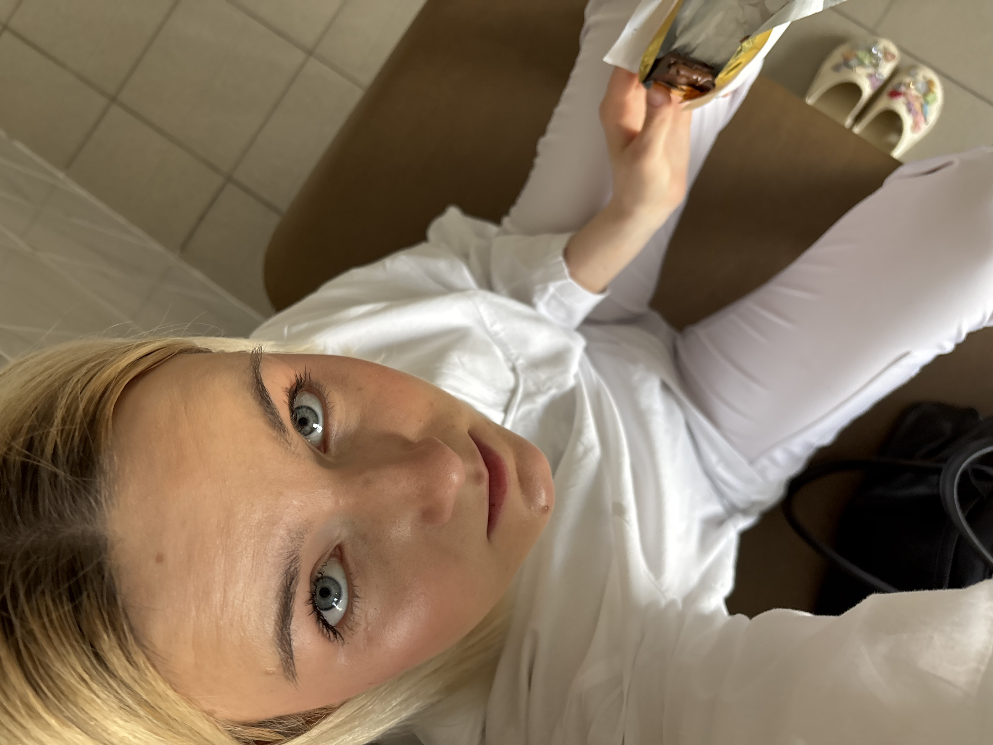 A young woman with blonde hair and striking blue eyes is the central subject of this indoor, high-angle shot, looking directly up at the camera. She appears to be in her 20s or 30s. Her hair is parted down the middle, framing her face, and her eyebrows are well-defined. Her wide, attentive eyes are a prominent feature, reflecting natural light.
She is dressed in a crisp white long-sleeved shirt and matching white pants, suggesting a light and casual attire. Her left hand, visible in the upper right portion of the frame, is holding an unwrapped chocolate bar, with a portion of the chocolate visible within the metallic foil wrapper. She seems to be in the process of eating or about to eat it.
The setting appears to be a room with light-colored square floor tiles. To her right, a brown surface, possibly an armrest of a couch or a low table, is visible. On the floor next to this brown surface, a pair of light-colored, embellished slippers with colorful designs (resembling Crocs or similar comfort footwear) are neatly placed. A dark-colored bag is partially visible near her hip on the left side of the frame. The overall impression is one of a relaxed, casual moment captured from an overhead perspective, likely in a home or private space within Saint Petersburg, Russia. No visible text is discernible in the image.