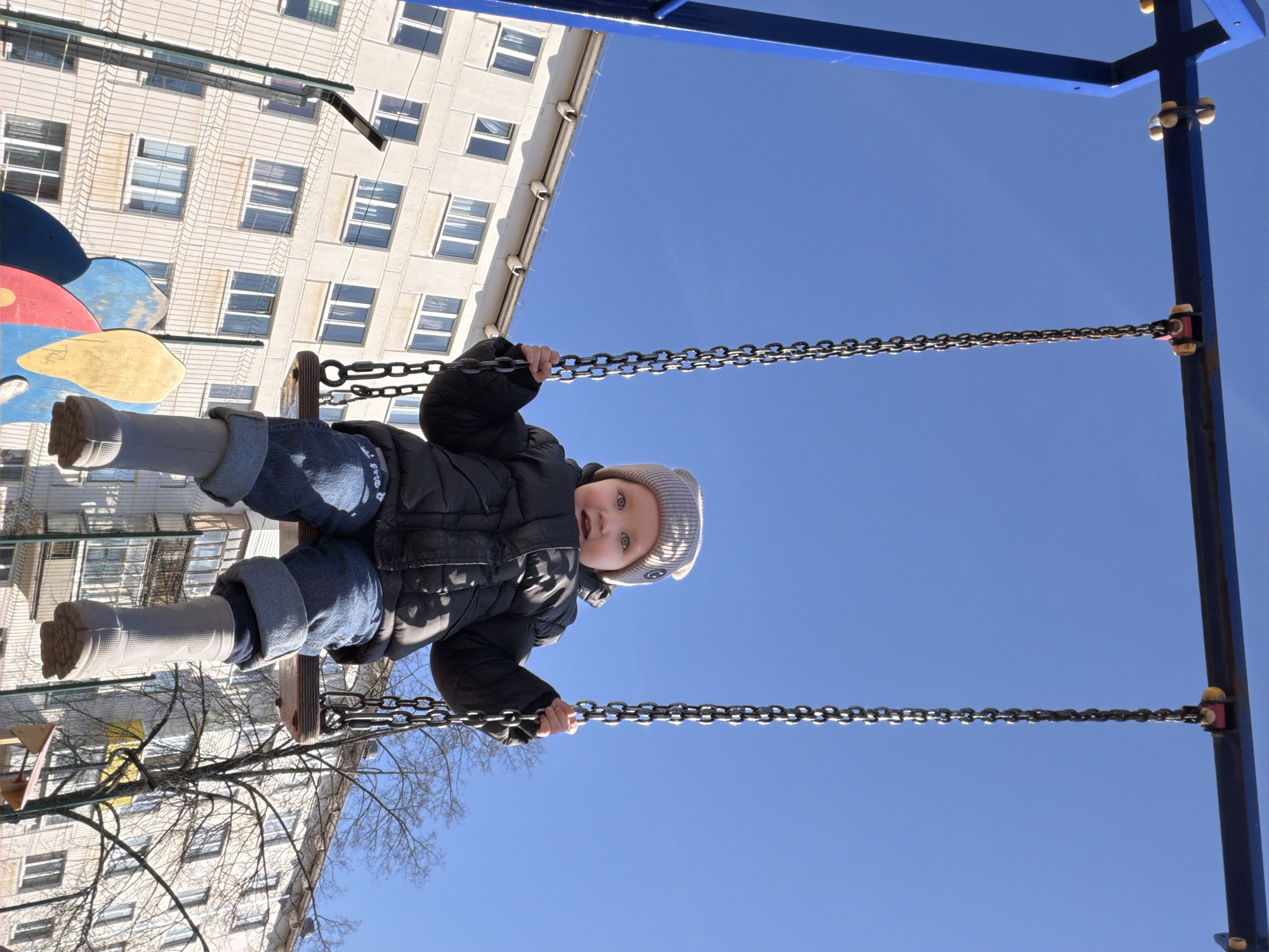 A young child, likely a toddler, is captured mid-swing on a bright, clear day, looking directly at the viewer with wide, curious eyes. The child is dressed for cool weather in a black puffy jacket, blue jeans, and a light grey knitted beanie adorned with a small emblem. Light-colored boots or thick socks cover their feet, while their hands firmly grip the metal chains of the swing set.
The scene is an outdoor playground, with the child positioned against a stark, vibrant blue sky that occupies most of the background. To the left, a large, light-colored multi-story apartment building with numerous rectangular windows provides an urban or residential backdrop. Visible drainpipes run down the building's facade. Some bare tree branches are also present, suggesting the season is late winter or early spring. Partially visible behind the child's legs is a colorful piece of playground equipment, possibly a flower-shaped structure in shades of red, yellow, and blue. The sunlight is bright, creating clear highlights on the swing chains and the child's face. The moment conveys a sense of innocent joy and curiosity outdoors in Chaikovsky, Russia.