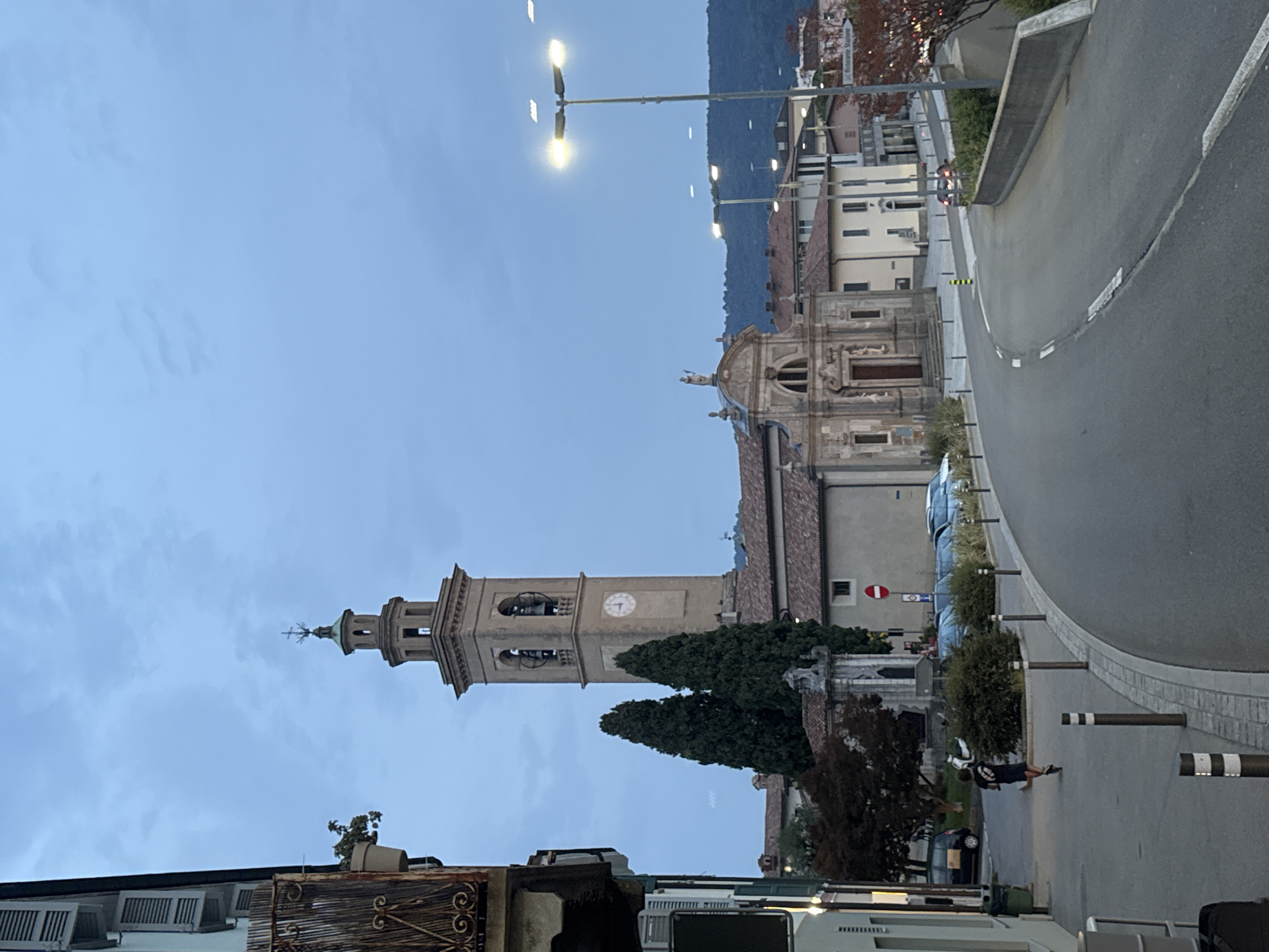 The image captures a street scene in Balerna, Switzerland, dominated by a tall church bell tower. The tower, made of stone, has multiple levels and features a clock face and visible bells. Adjacent to the tower is the church, a historical building with an arched entrance. A road curves through the scene, with parked cars and streetlights visible. A person is seen riding a scooter on the sidewalk. The sky is overcast, indicating twilight or a cloudy day. The overall environment suggests a European town with historical architecture.