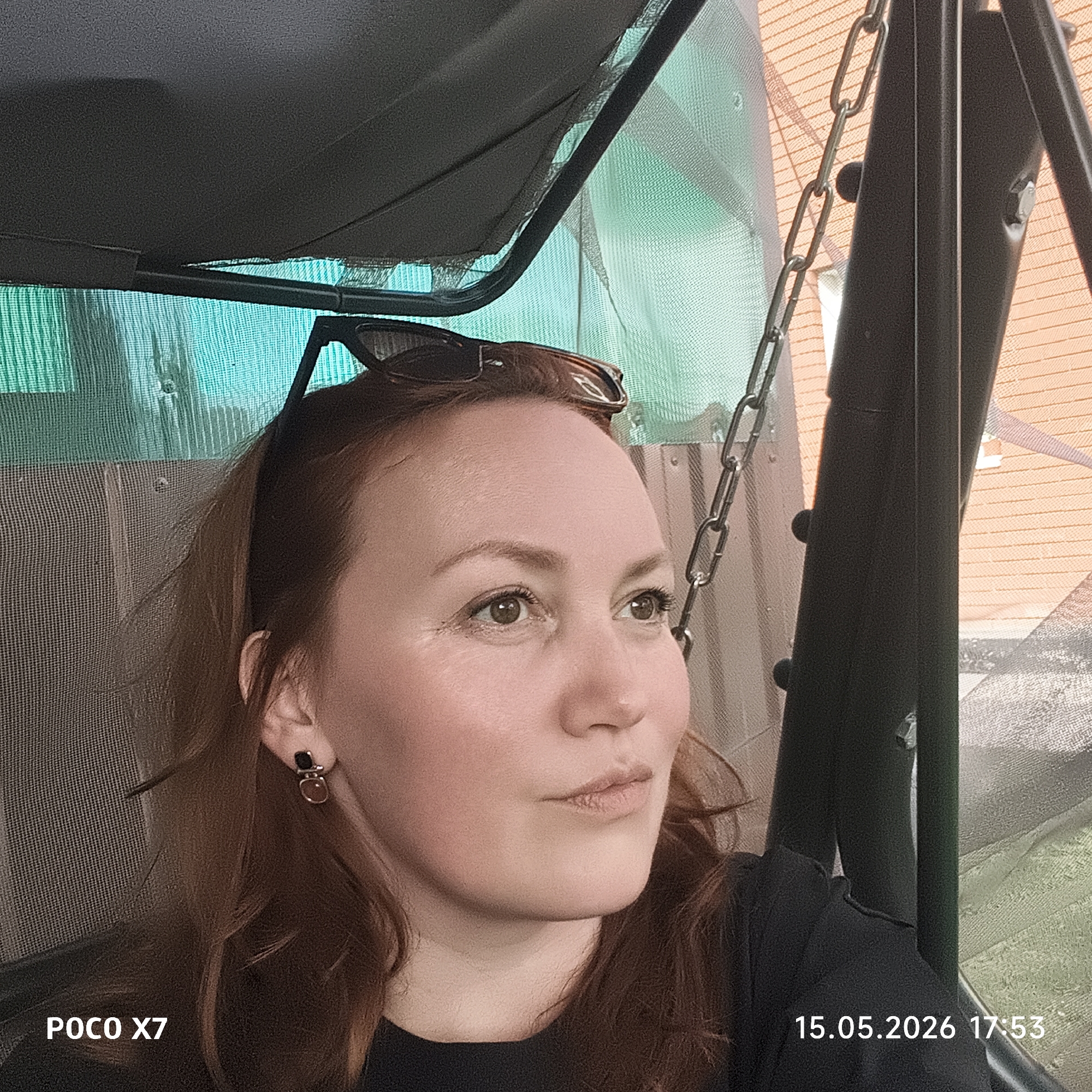 A medium indoor/outdoor shot depicts a woman seated, likely in a swing or covered patio area, with elements suggesting both an enclosed space and an exterior setting. The woman is fair-skinned with reddish-brown hair and is wearing dark clothing and earrings. Sunglasses are perched atop her head. The background features a translucent green screen or netting, along with metal supports and chains indicating a swing-like structure. There is also a glimpse of a building with brick or siding in the background. The image contains a timestamp indicating 17:53 on 15.05.2026 and "POCO X7" in the lower left corner, indicating the device used to capture the image. There is no information to determine the image's location being Chaikovsky, Russia.