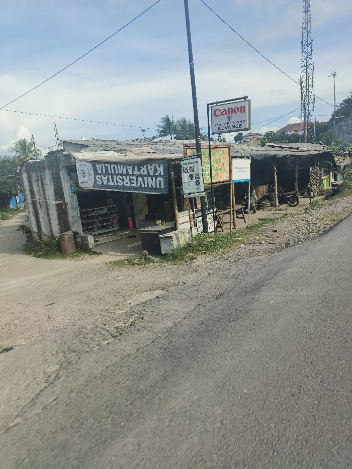 The image shows a street scene in Sukatani, Indonesia (latitude: -6.62, longitude: 107.405). In the foreground, a paved road with a dirt shoulder leads toward a small storefront with a sign that reads "UNIVERSITAS KARTAMULIA." The storefront is a partially enclosed structure with a corrugated metal roof. A sign advertising "Canon ImageRUNNER ADVANCE" is mounted on a metal pole. The sign is partially obscured by trees. Several other signs advertising businesses are visible. The storefront is partially obscured by trees and shrubs. An upright metallic radio tower is visible in the background. There are no people or vehicles visible in the scene. The sky is clear and blue. The scene appears to be a rural commercial area.