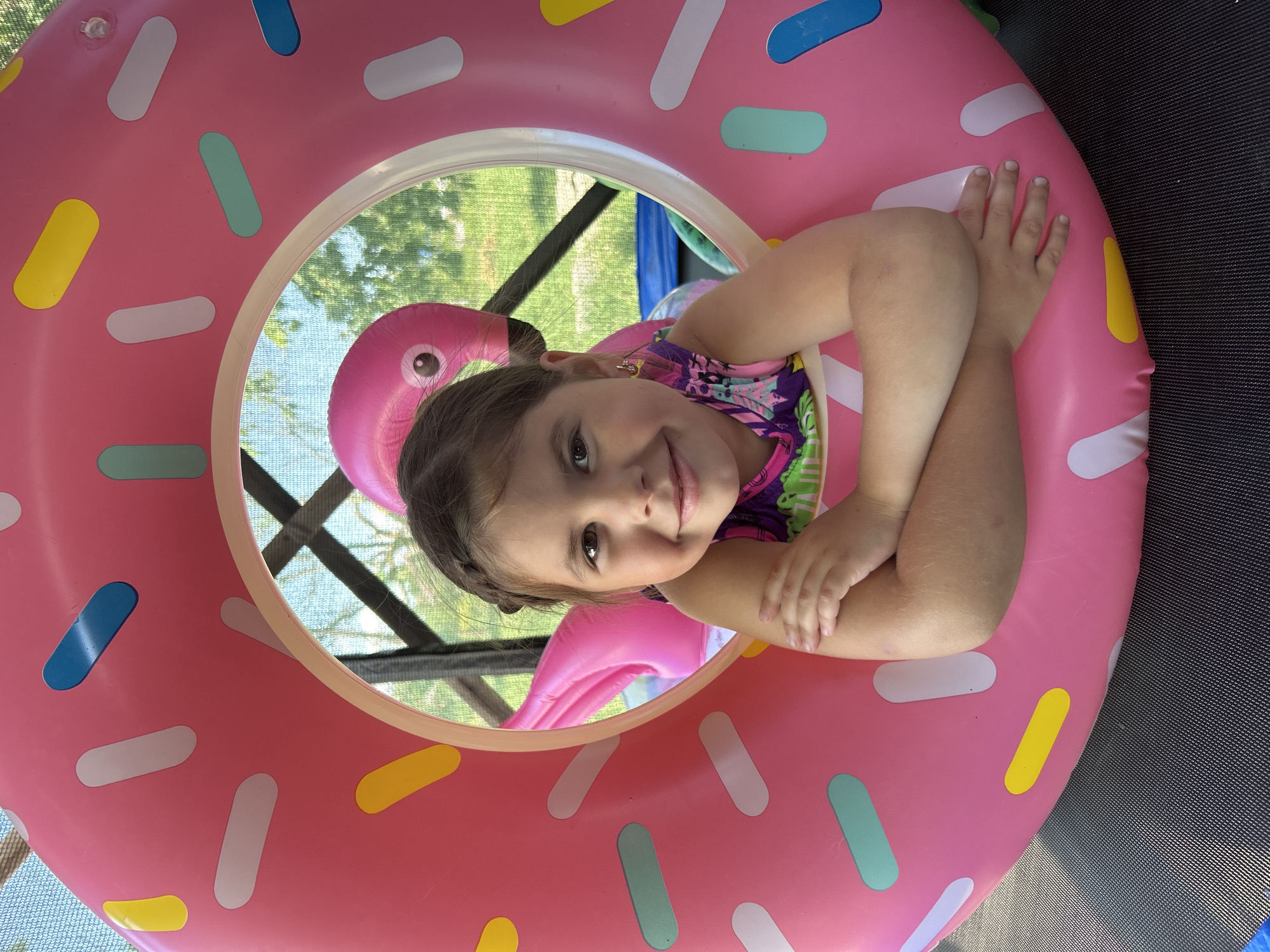 A young girl with brown hair, wearing a colorful swimsuit, smiles as she lies on her side within a large, pink inflatable donut. The donut has white, yellow, blue, and green sprinkles. Behind the girl, a pink inflatable flamingo can be seen. The background shows greenery and a dark, possibly netted structure, suggesting an outdoor setting. The girl's arms are crossed over her chest, and her hands are visible. The lighting indicates it is daytime. The girl appears happy and relaxed.