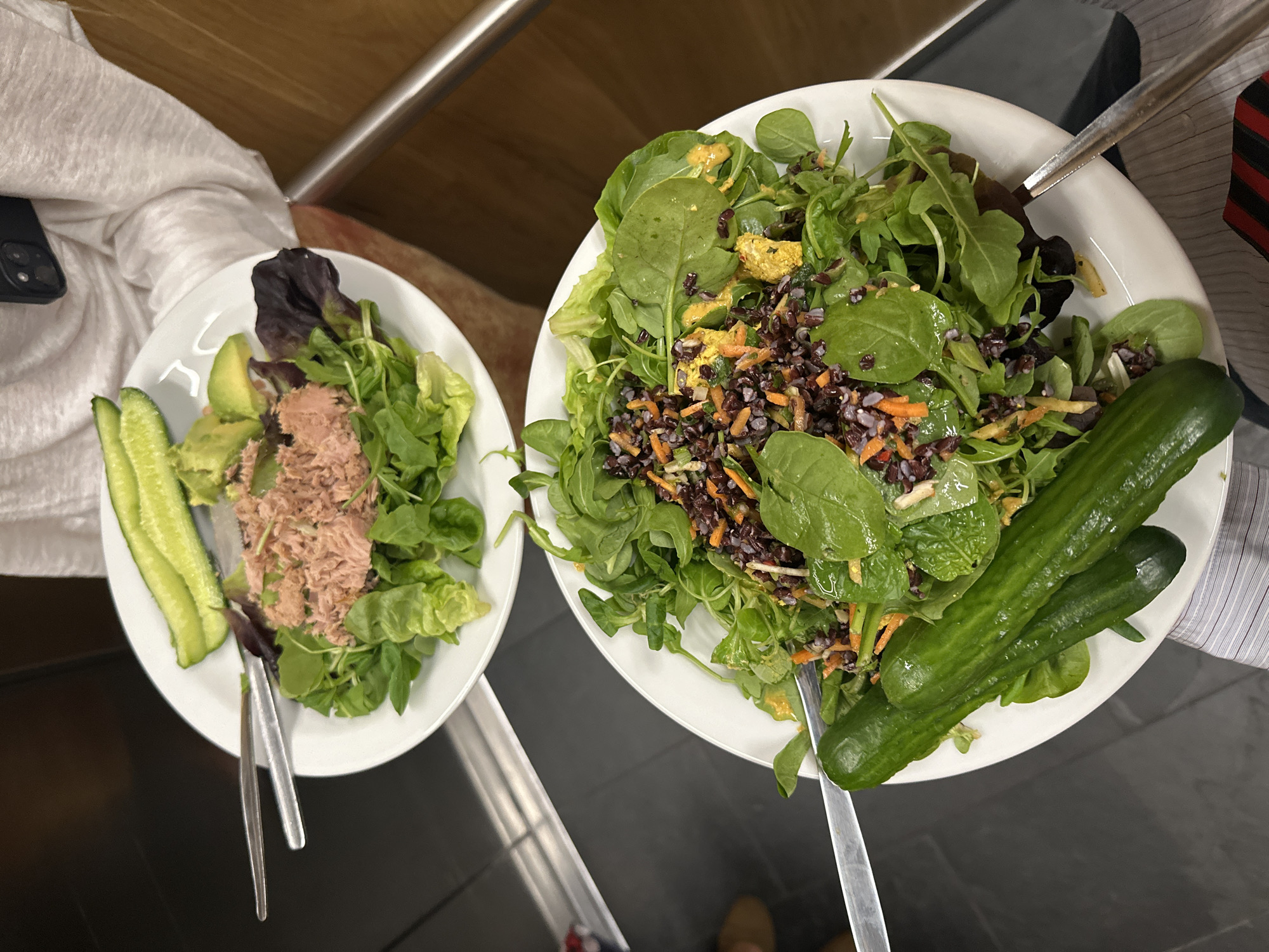 Two white plates hold salads. The plate on the left contains lettuce, avocado slices, tuna, and cucumber slices. The plate on the right contains a salad of mixed greens, black rice, carrots, yellow peppers, cucumbers, and salad dressing. Each plate has utensils. The scene is indoors.