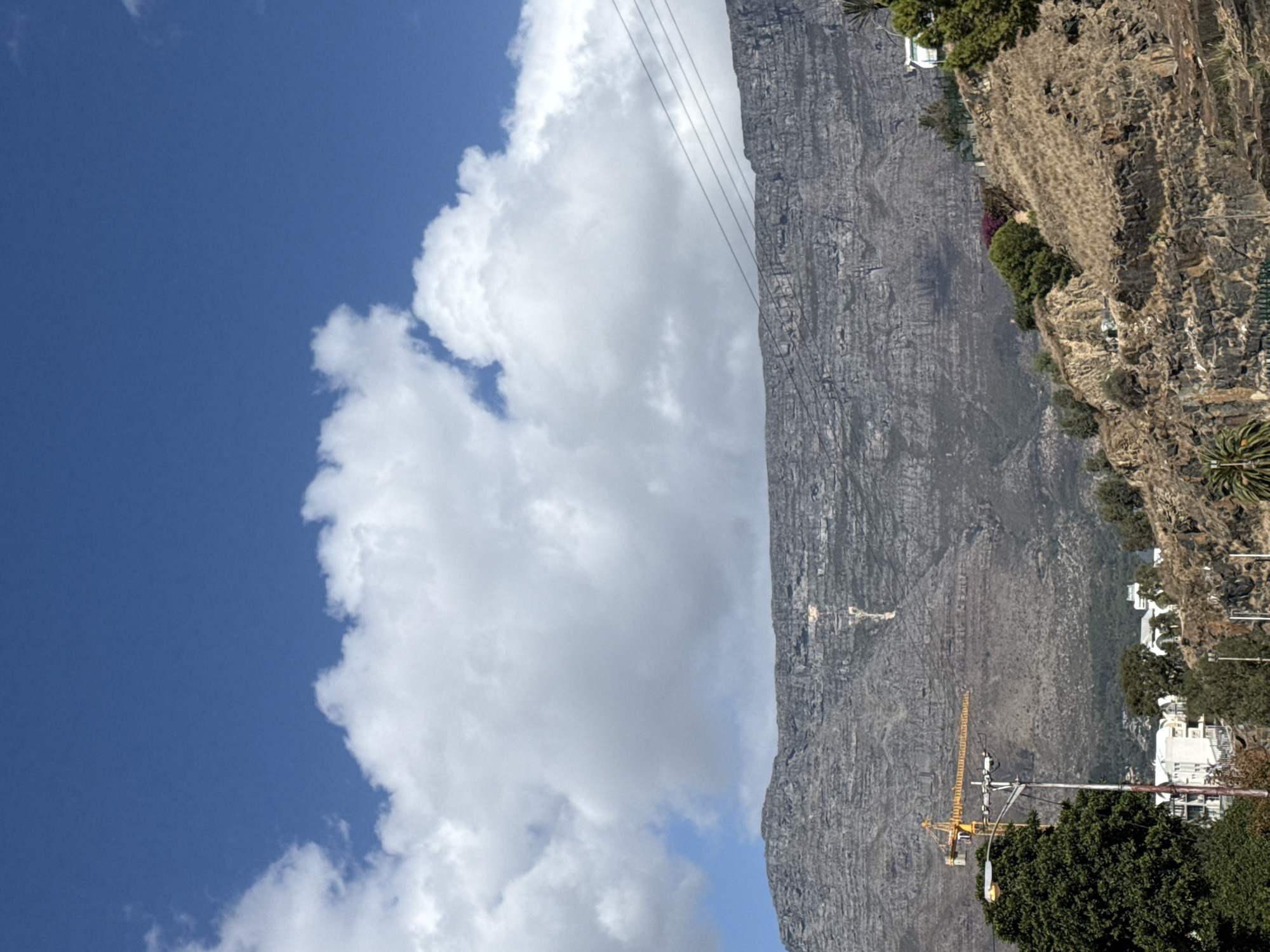 The image shows Table Mountain in Cape Town, South Africa, on a sunny day with blue sky and large white clouds to the left. The mountain's rocky face is grey, textured with vertical lines. Near the base, a yellow construction crane is visible. Houses and trees are on the right side of the mountain. Power lines run along the top of the mountain.