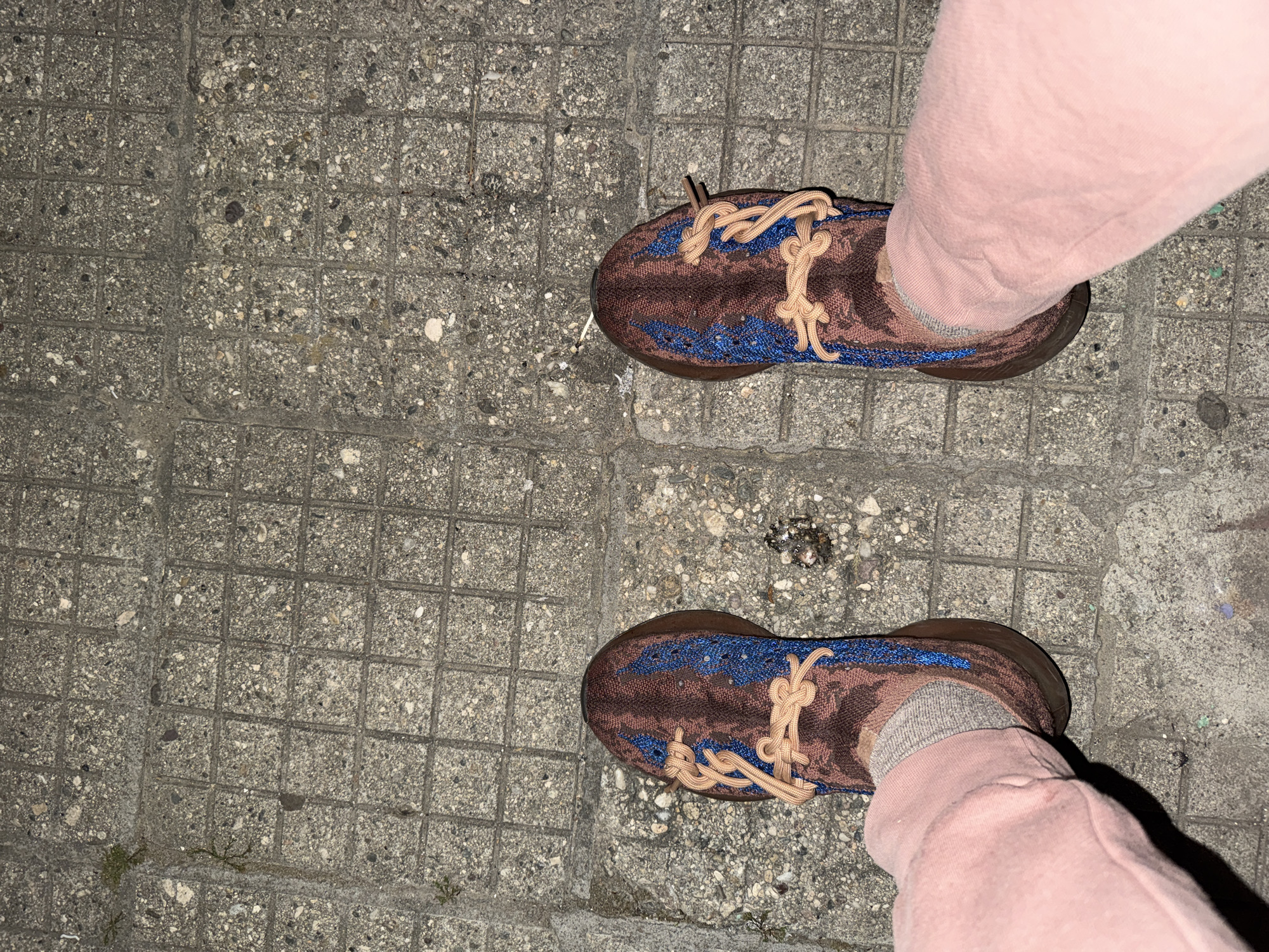 The image shows a person's feet wearing brown and blue sneakers with light brown laces and light pink sweatpants, standing on a sidewalk composed of square concrete blocks. The sidewalk appears to be slightly weathered and stained in some areas. The picture has been taken from an elevated perspective, looking down at the feet. The scene does not provide enough details to discern the specific location within the city of Polena, Bulgaria. No interactions or actions are visibly occurring.