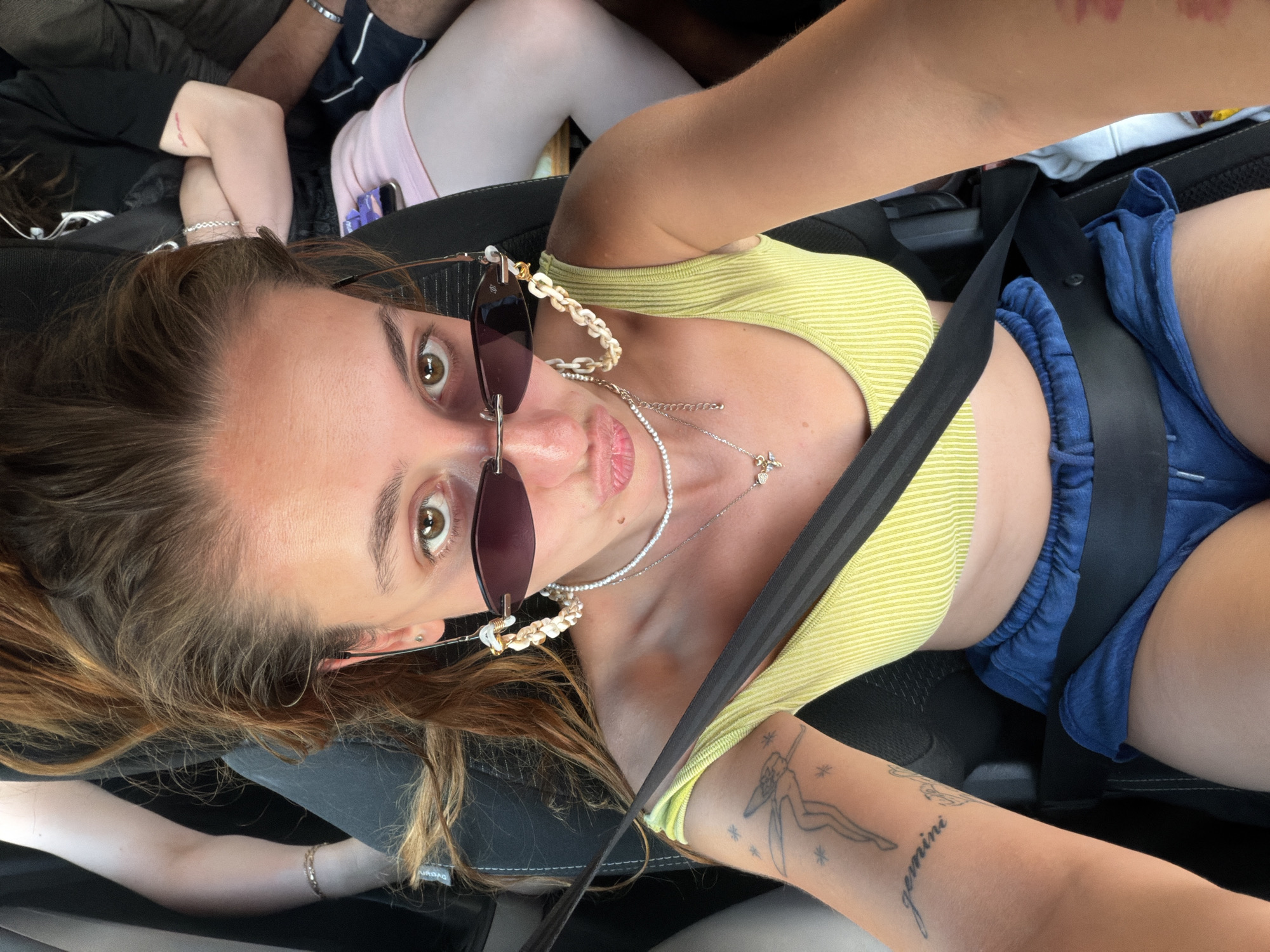 A young woman with long, auburn hair is taking a selfie while sitting in the passenger seat of a vehicle. She is wearing a lime-green tank top, blue shorts, and sunglasses perched on her nose. She is also wearing a pearl necklace and a decorative chain attached to her sunglasses. A seatbelt is across her chest. Her left arm is visible, showing a tattoo of a winged figure and the word "gemini." Other people are visible in the background, seated in the vehicle. The image appears to be taken during the day, with natural light illuminating the scene. The location is not specified in the image or the provided crops.