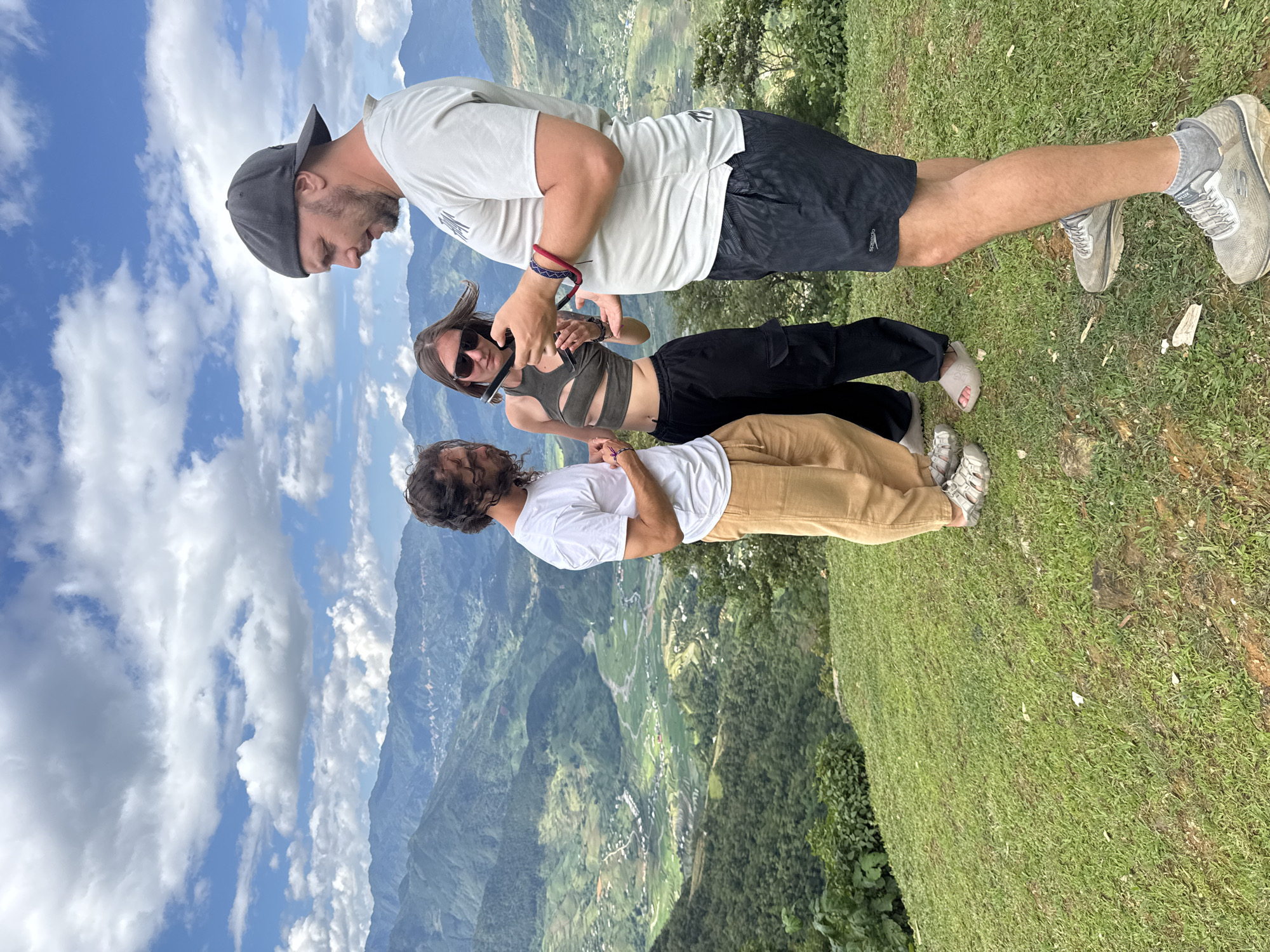 Three individuals are positioned on a grassy hillside overlooking a mountainous landscape in the city of Tu Le, Vietnam, under a partly cloudy blue sky.
The individual on the left, an adult male with dark curly hair and a beard, wears a white short-sleeved t-shirt, light brown loose-fitting pants, and light-colored open-toed sandals. He stands with his hands clasped in front, directing his gaze towards the central female and the object she holds. A red and blue braided bracelet is visible on his left wrist.
The central individual, an adult female with dark hair, wears sunglasses, a dark olive-colored crop top with cutouts, and dark cargo-style pants. Her footwear consists of light grey slip-on sandals. She holds a small, dark, elongated object with both hands, manipulating or examining it intently while looking down. A dark bracelet is visible on her right wrist.
The individual on the right, an adult male with a beard and a dark baseball cap worn backward, wears a light grey short-sleeved t-shirt with a subtle graphic and dark short-style swim trunks with "Speedo" branding visible on the left leg. He wears grey athletic sneakers with grey socks. He stands slightly turned, observing the object held by the female, with his hands positioned as if engaged in the shared focus. He also wears a red and blue braided bracelet on his left wrist.
The background reveals a vast mountainous terrain with multiple green peaks receding into the distance. Below the viewpoint is a deep valley characterized by lush green vegetation, numerous terraced fields, scattered small buildings indicative of human settlements, and a winding river or stream. The foreground consists of short, green grass with visible patches of earth and small rocks. The sky features scattered white cumulus clouds against a clear blue expanse. The three individuals appear to be focused on the object held by the female, suggesting a shared point of interest or activity.